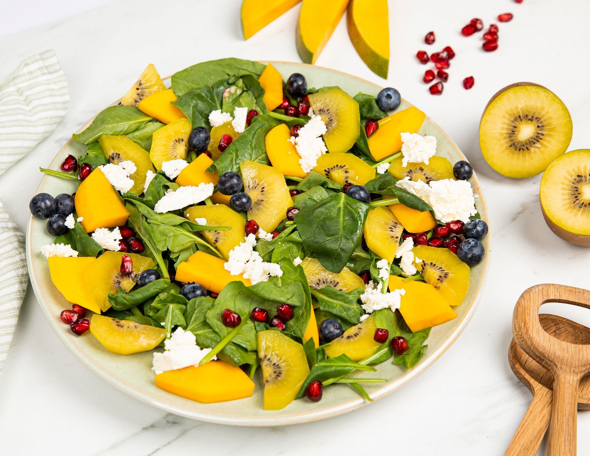 Salat med spinat, mango, kiwi, blåbær, granateplekjerner og smuldret hvitost, servert på et fat med frisk frukt og salatbestikk ved siden av.