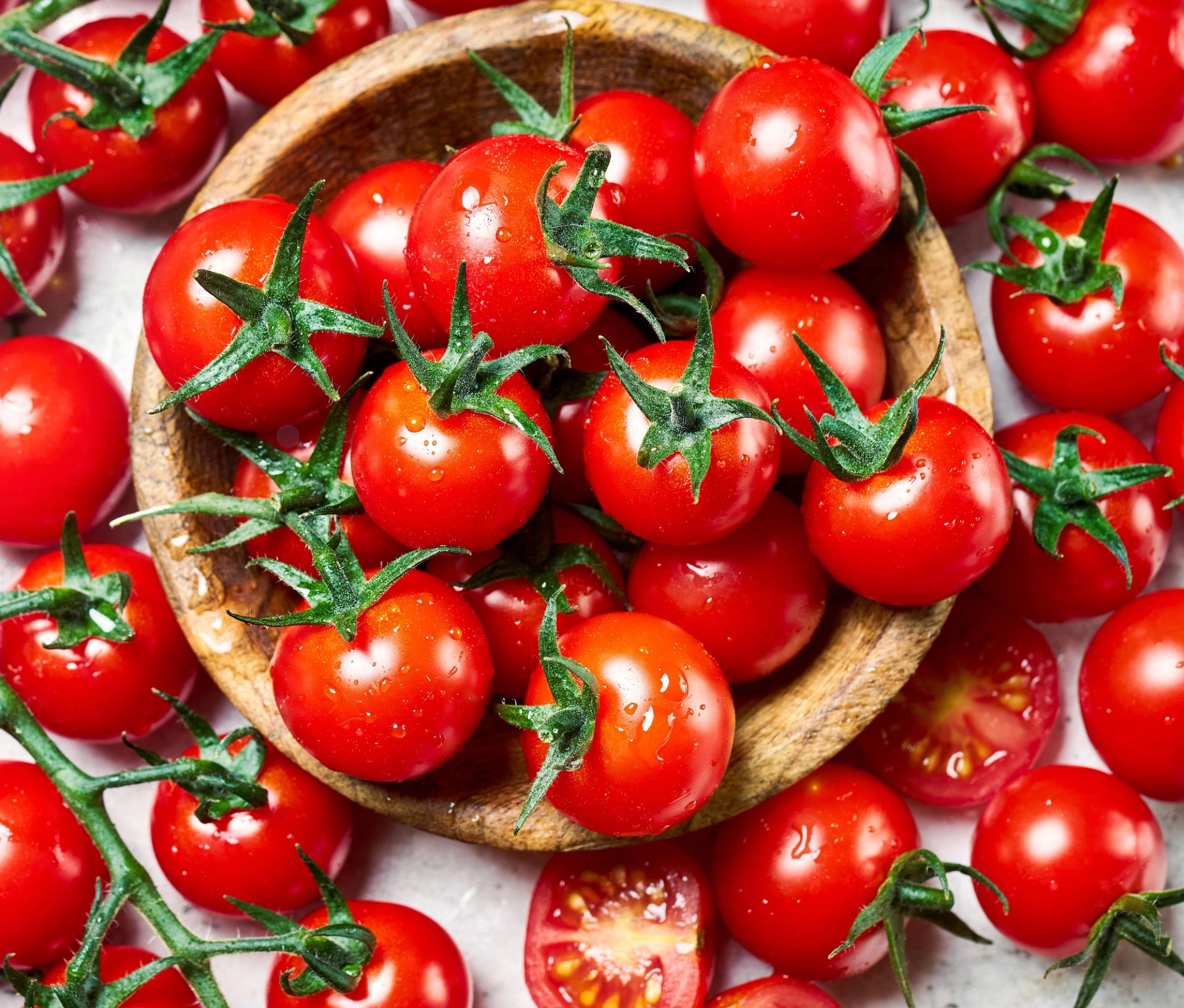 En trebolle fylt med røde cherrytomater med grønne stilker, omgitt av flere cherrytomater og noen halve tomater spredt utover en lys bakgrunn.
