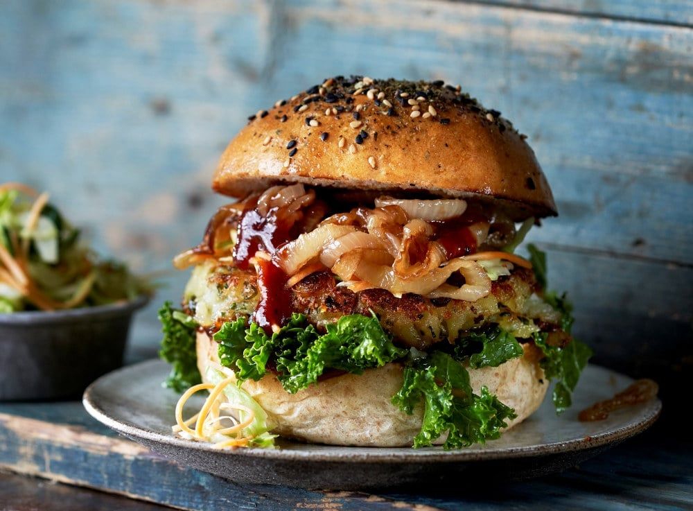 Burger på tallerken med sesamdekket briochebrød, saftig burger, karamellisert løk, grønnkål og saus, fotografert mot en rustikk blå trebakgrunn.