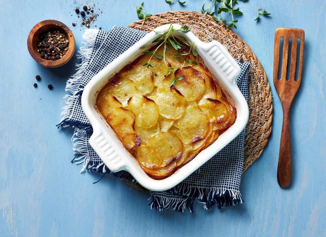 Ovnsbakt Pommes Anna med lagdelte potetskiver i en ildfast form, gyllen og sprø på toppen, servert på et blått bord med pepper, hvitløk og timian ved siden av.