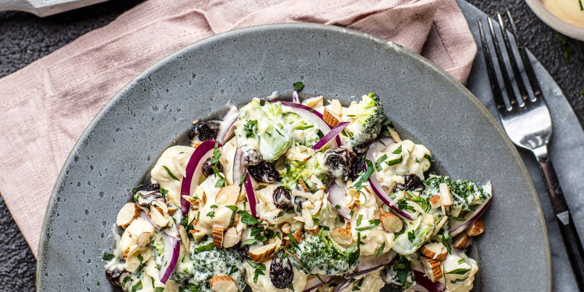 Brokkoli‑ og blomkålsalat med kremet dressing, rødløk, rosiner og hakkede nøtter, servert på tallerken.