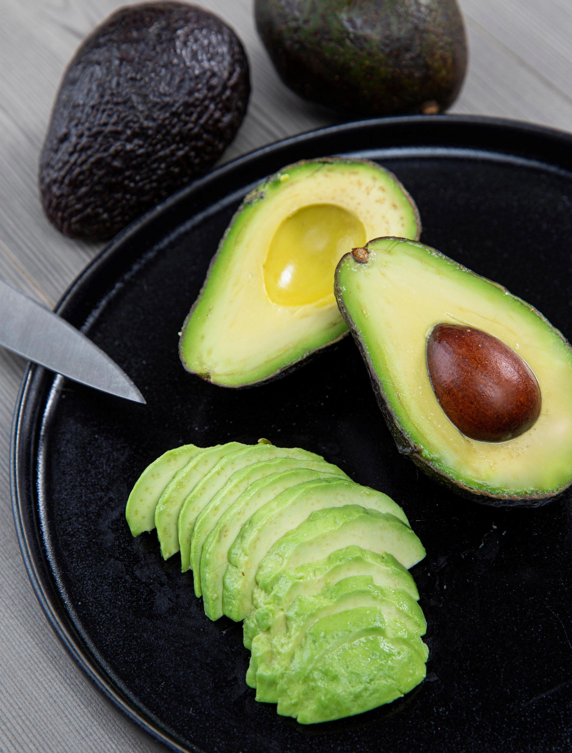 Et svart fat med avokado: én halv avokado med stein, én uten stein, og flere tynne avokadoskiver foran, med hele avokadoer og en kniv i bakgrunnen.