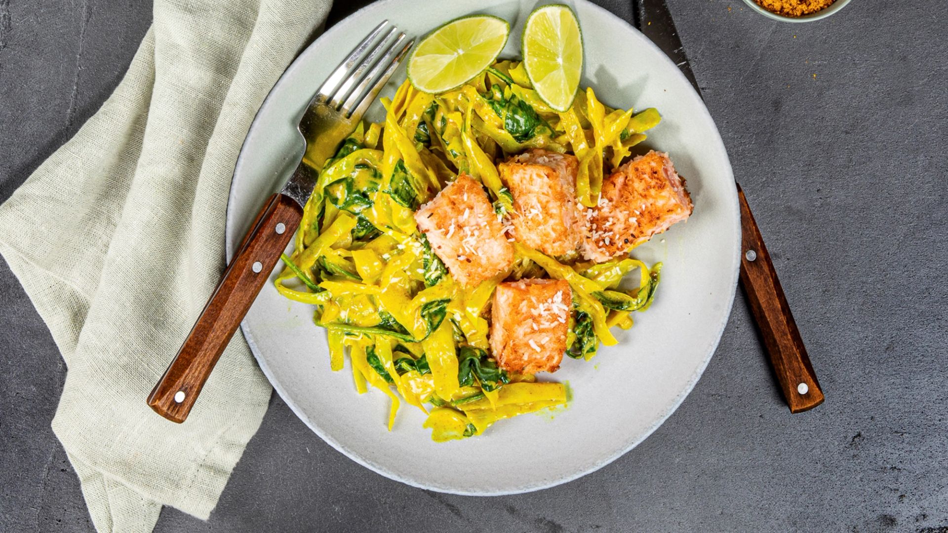 Et bilde sett ovenfra av en tallerken med fire stykker kokosstekt laks servert på en seng av gul currykål og grønn spinat. Retten er toppet med to limebåter og et dryss av kokos. 
