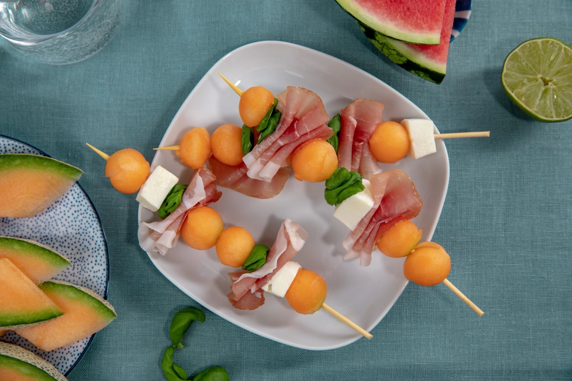 Tre små trespyd med kuler av oransje cantaloupemelon, tynne skiver spekeskinke, terninger av hvit ost og friske basilikumblader. Spydene ligger på en hvit tallerken på en blå tekstilduk, omgitt av biter med vannmelon, cantaloupe og lime.