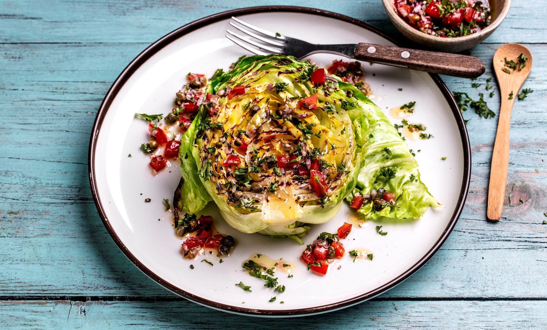 Et grillet kålskive servert på en hvit tallerken mot et lyseblått tregulv. Den grillede kålen er toppet med en frisk salsa av hakkede tomater, løk og kapers, samt et dryss av finhakket urter..