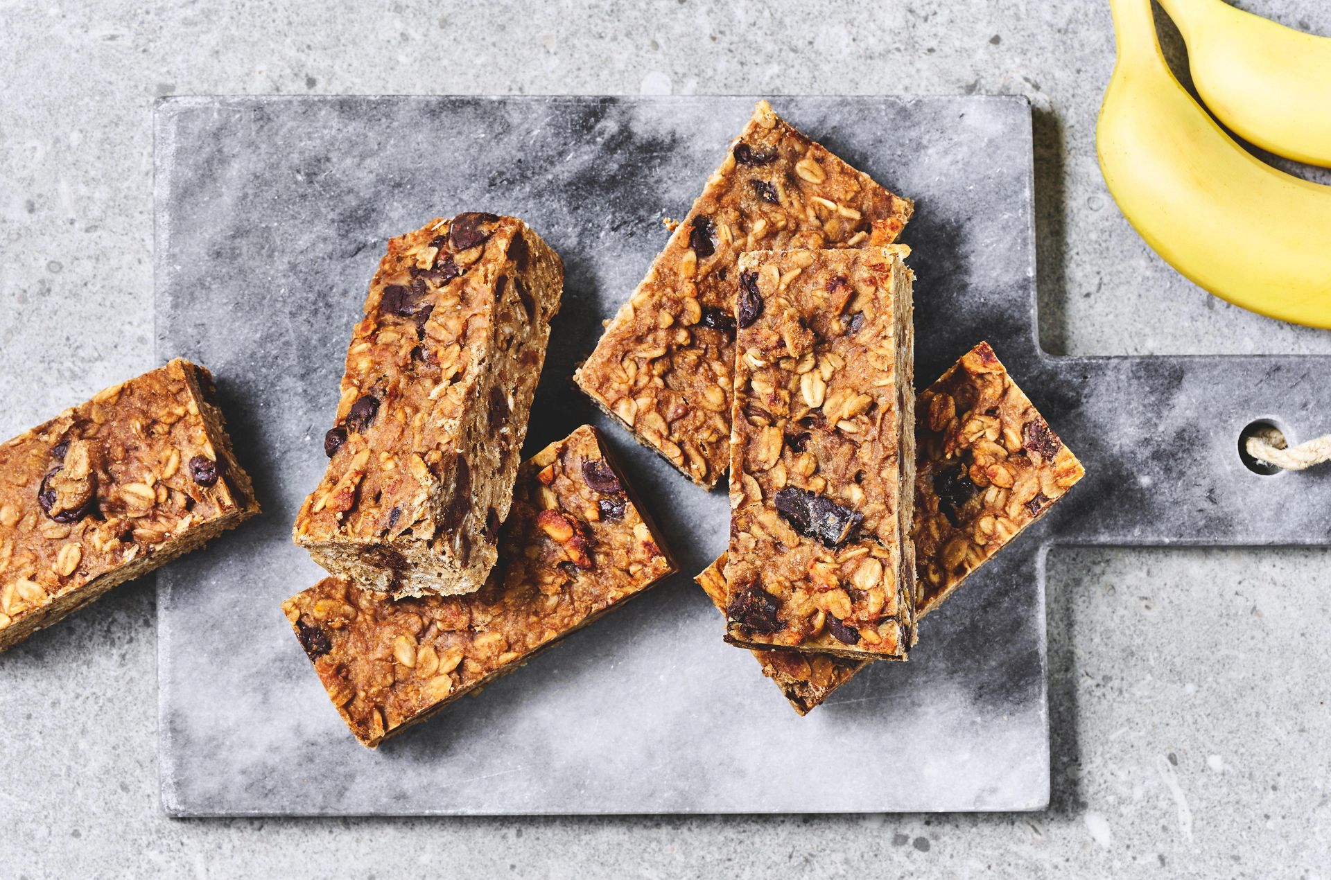 Hjemmelagde müslibarer med havregryn og biter av tørket frukt ligger stablet på et grått steinbrett. Ved siden av brettet ligger en klase bananer på en lys grå benkeplate. Bildet er tatt ovenfra i naturlig lys.