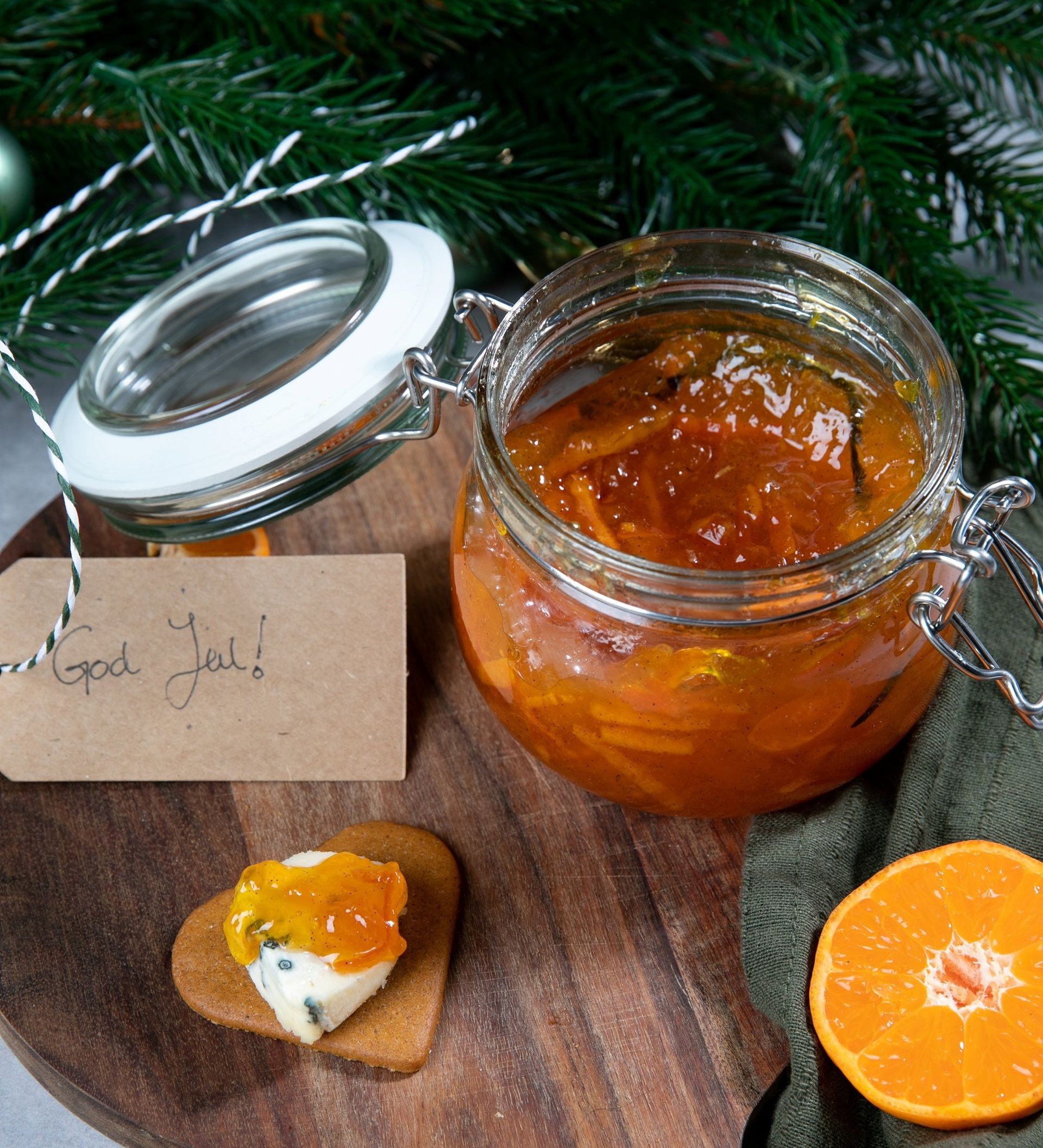 Julens klementinmarmelade med synlige klementinstrimler, i glass stående på en trefjøl pyntet med granbar.
