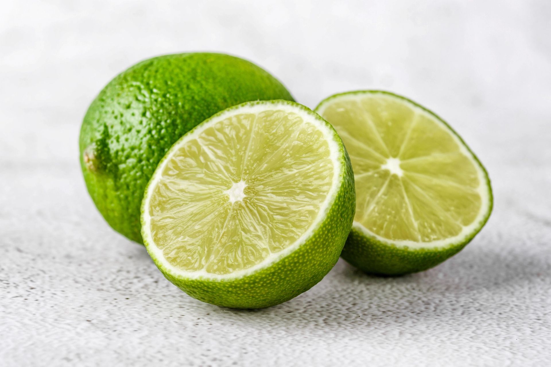 En hel lime og to halve lime plassert på en lys overflate, der de delte limefruktene viser den lysegrønne, saftige fruktkjøttsiden.