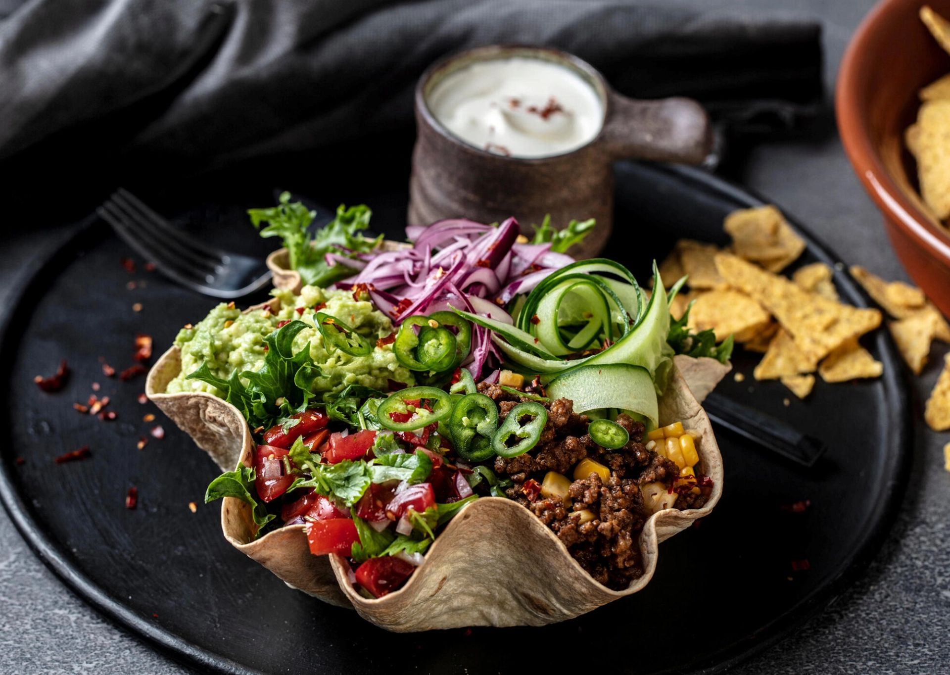 Tortillaskål fylt med tacofyll, guacamole, frisk salat, tomat, mais, rødløk, agurkstrimler og skivet jalapeño, servert på et mørkt fat med nachochips i bakgrunnen.