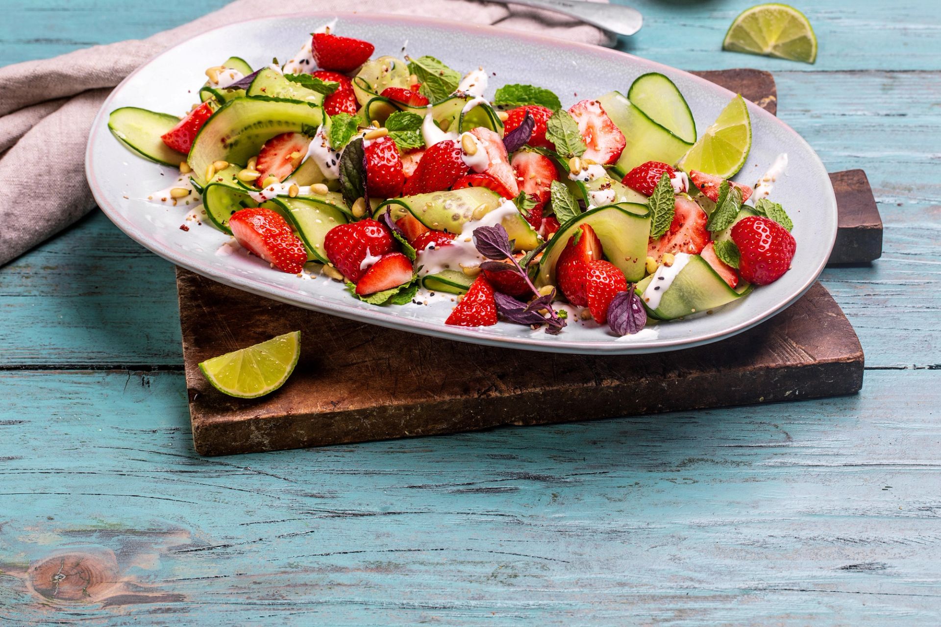 Frisk agurk- og jordbærsalat med basilikum, mynte, pinjekjerner og yoghurt servert på fat med limebåter.