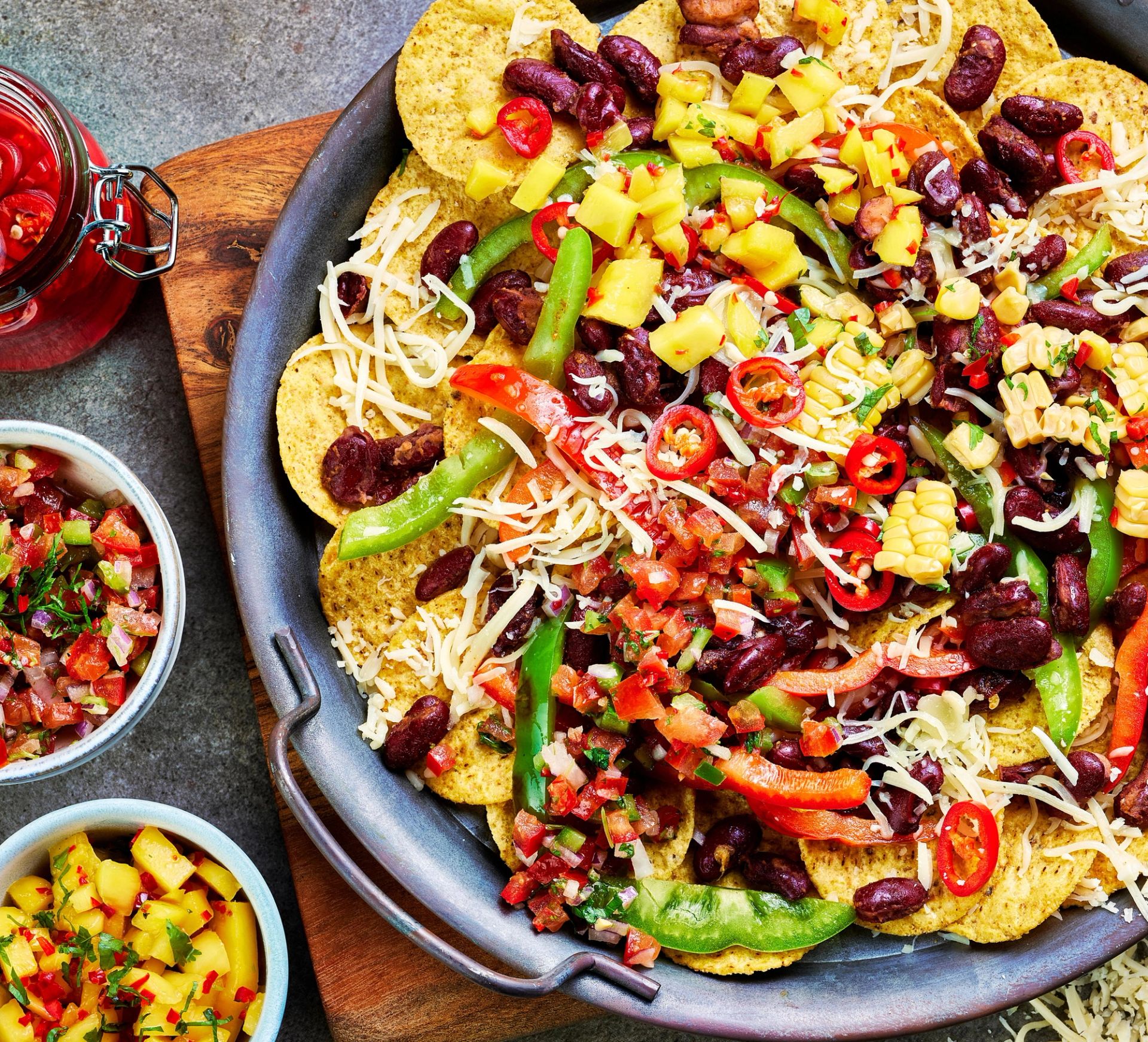 Et stort fat med nachos toppet med revet ost, røde kidneybønner, mais, grønn paprika, rød chili og frisk salsa. Rundt fatet står små skåler med mango, pico de gallo og syltet chili, på et rustikt bord.