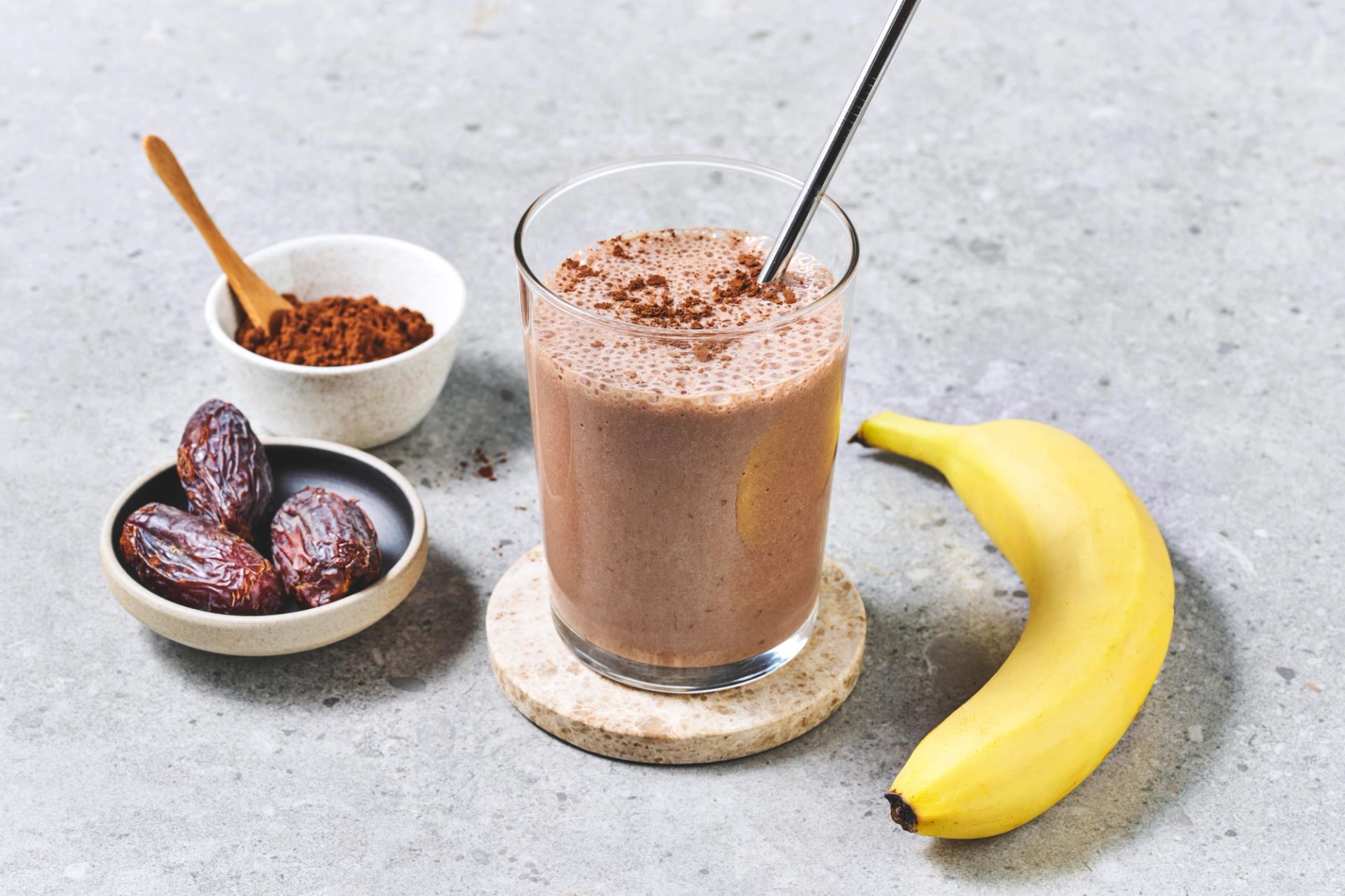 Et glass sjokoladefarget smoothie toppet med kakaopulver, servert med metallsugerør på en steinunderlag. Ved siden av står en skål med kakaopulver, dadler og en moden banan.