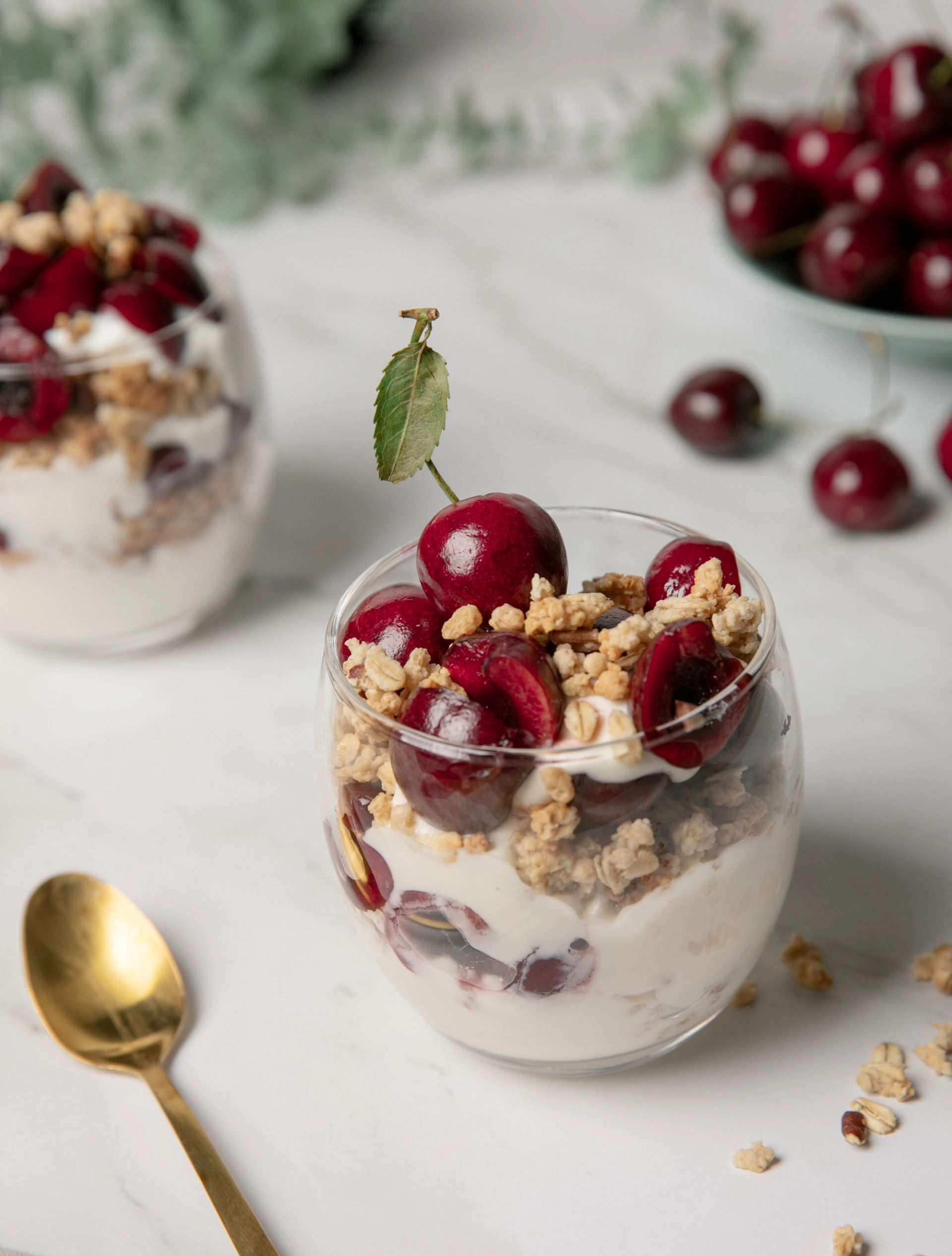 Et glass med yoghurt rørt med granola og halvdelte moreller, toppet med hele og delte mørkerøde moreller. I bakgrunnen står et lignende glass og en skål med moreller på et hvitt marmorlignende underlag. En gullfarget skje ligger ved siden av.