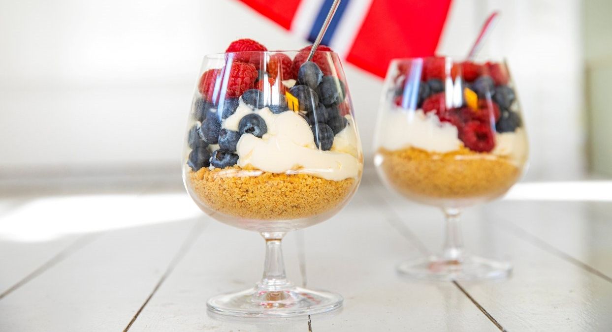 To porsjoner med ostekake på glass, fylt med smulebunn, kremet ostefyll og friske blåbær og bringebær står på et hvitt bord..