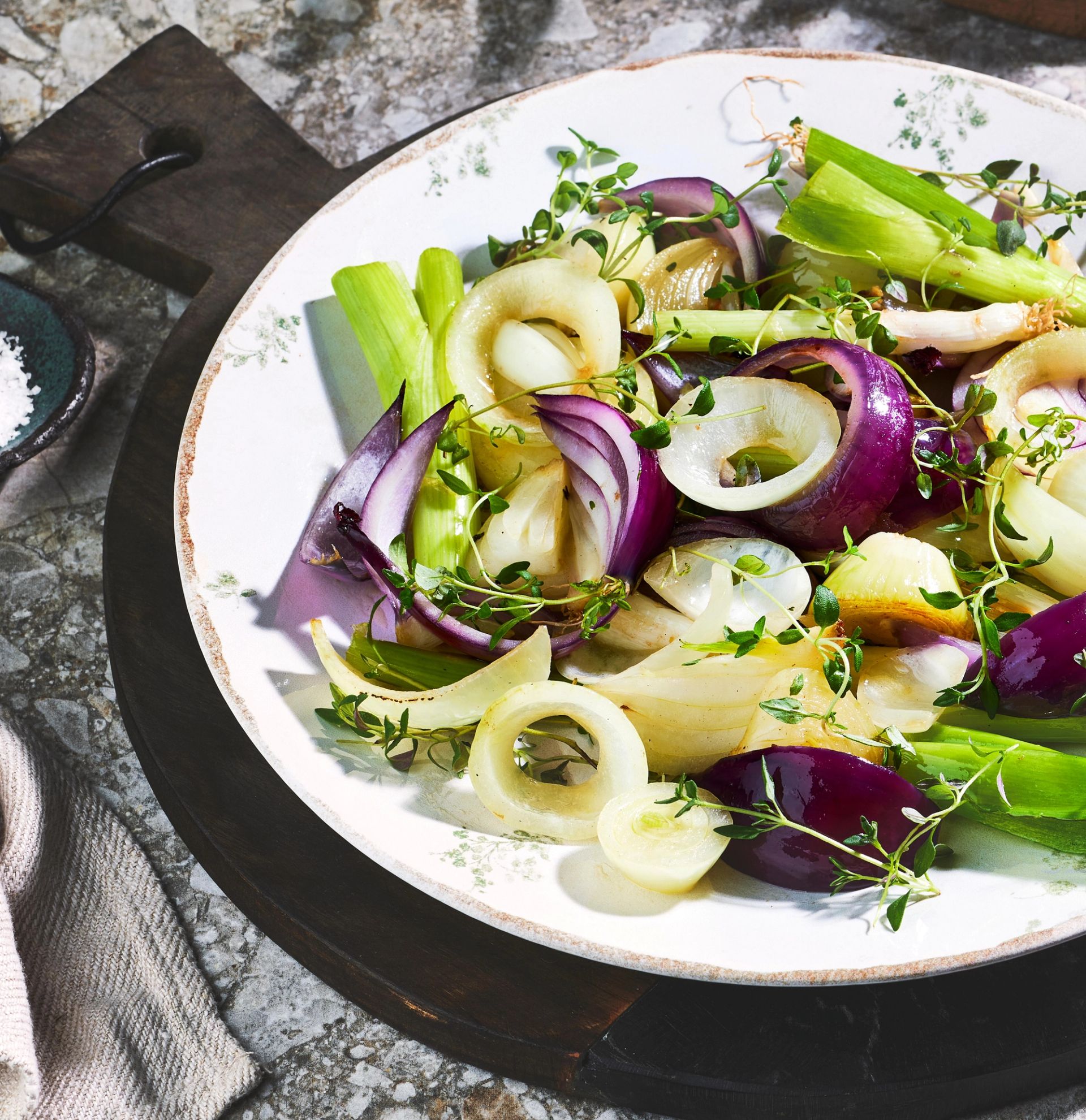 En stor tallerken fylt med en rustikk løksalat bestående av grillet purre i lange biter, rødløk i båter og lyse løkringer, toppet med friske urter. Grønnsakene har en blanding av myke og sprø teksturer, med lett karamelliserte og gyldne flater. Tallerkenen står på en mørk trefjøl med grovt salt og en steinoverflate i bakgrunnen, og lyset fremhever fargene og de naturlige strukturene i ingrediensene.