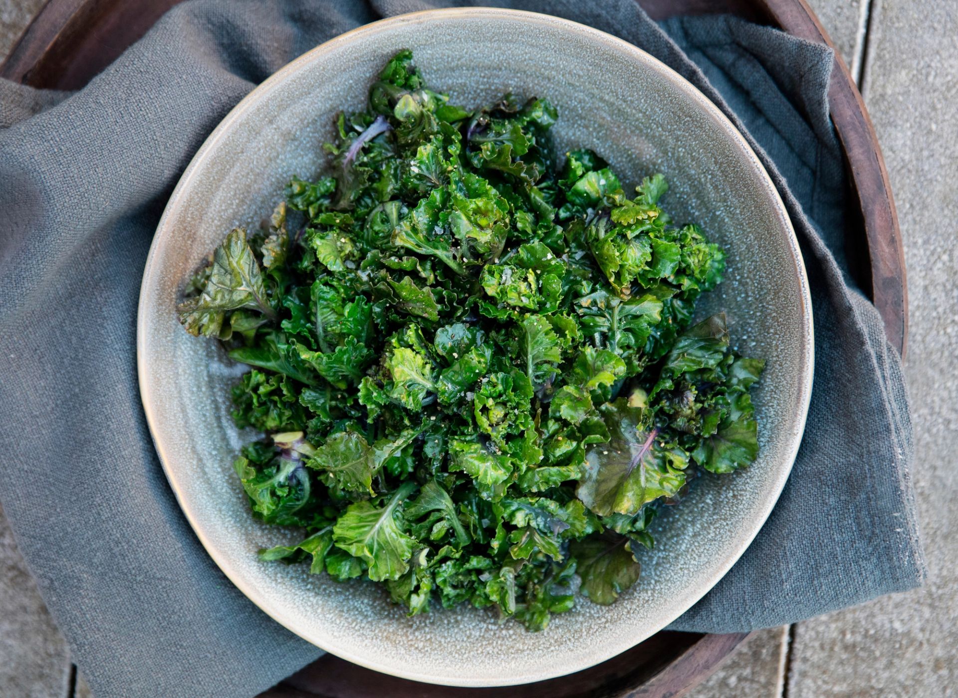 En skål fylt med frisk rosettkål sett ovenfra. De krusete, mørkegrønne bladene er vendt i en blank dressing. Salaten serveres i en lys keramikkbolle plassert på et mørkt tekstil, noe som fremhever de friske grønne fargene.