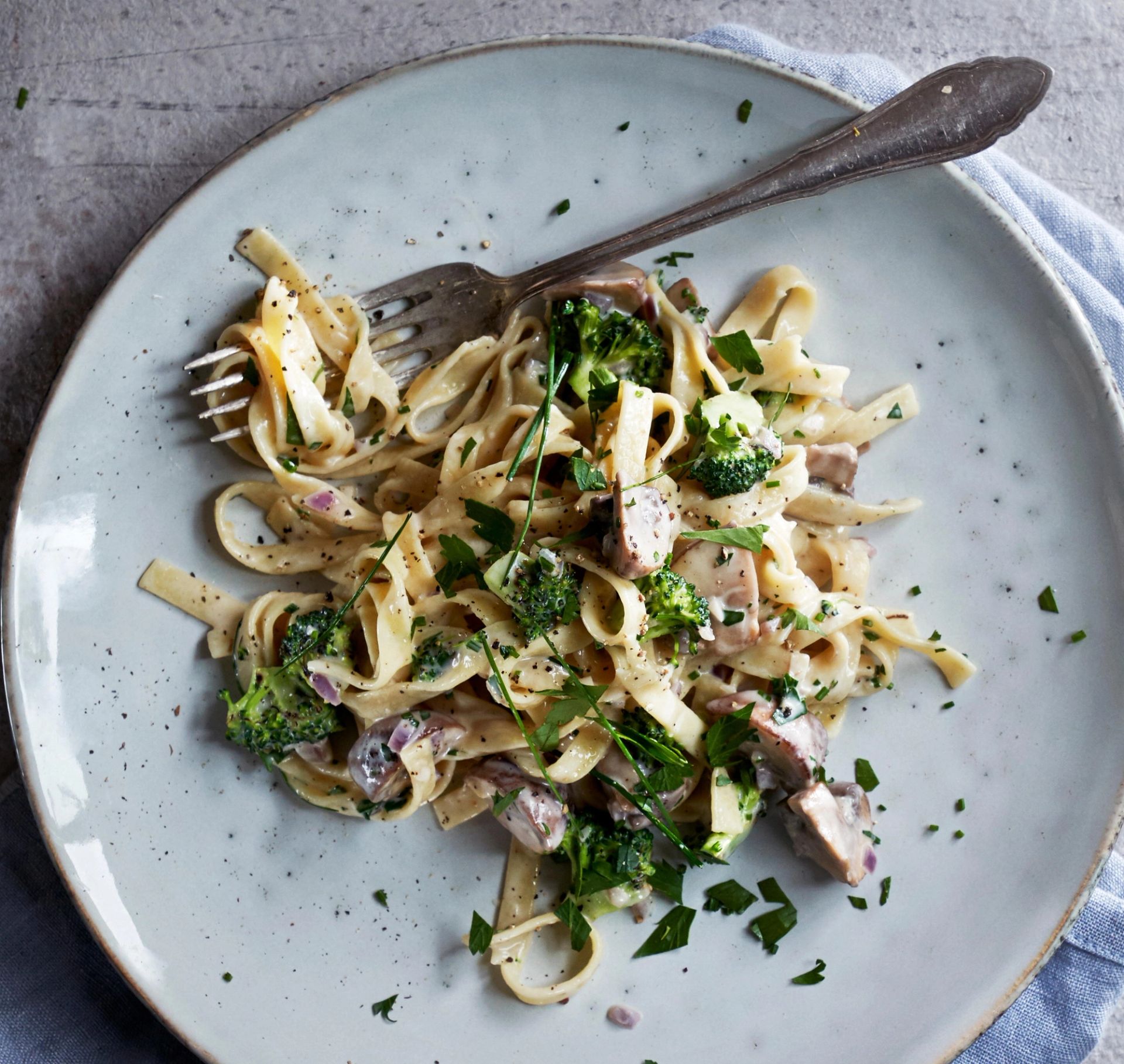 En tallerken med kremet pasta med sopp og brokkoli, servert med friske urter og nykvernet pepper, sett ovenfra på en lys keramisk tallerken med en gaffel ved siden av.