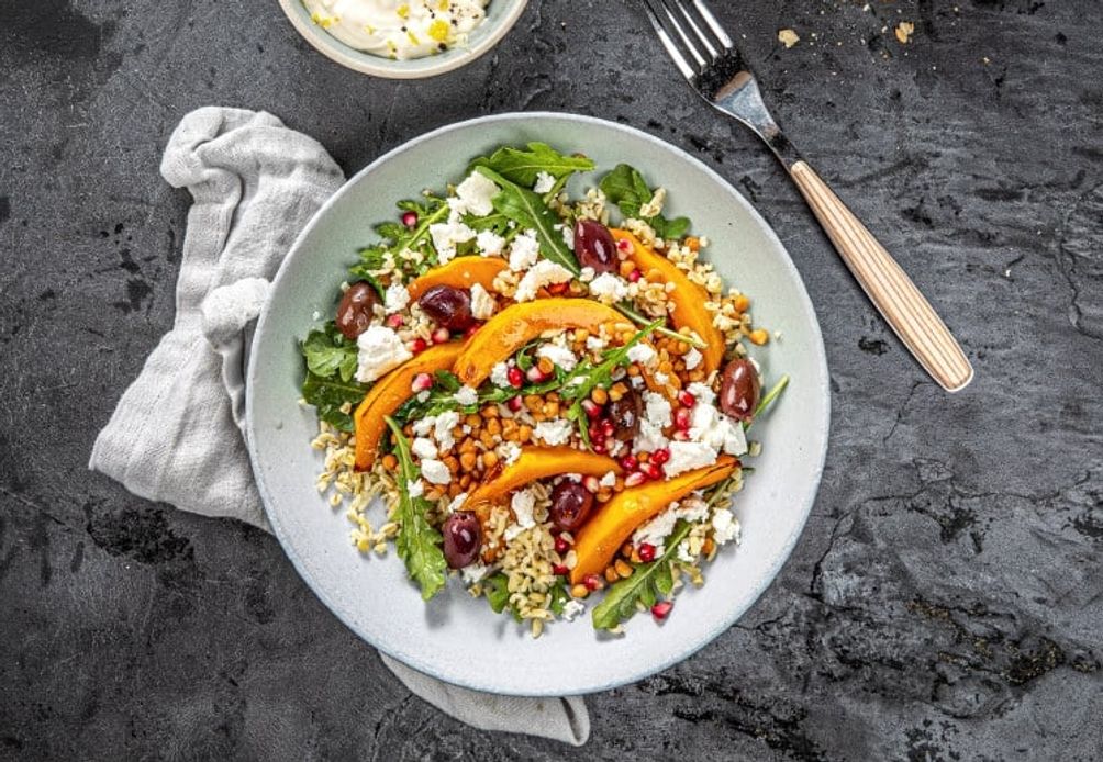 En fargerik bulgursalat servert i en lyseblå bolle på en mørkegrå, teksturert flate. Salaten består av kokt bulgur toppet med ovnsbakte skiver av gresskar, smuldret fetaost, svarte oliven, friske ruccolablader og røde granateplekjerner. 