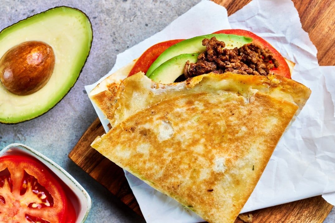 Sprøstekt quesadilla fylt med kjøttdeig og avokado, servert med skiver av tomat og delt avokado ved siden av.