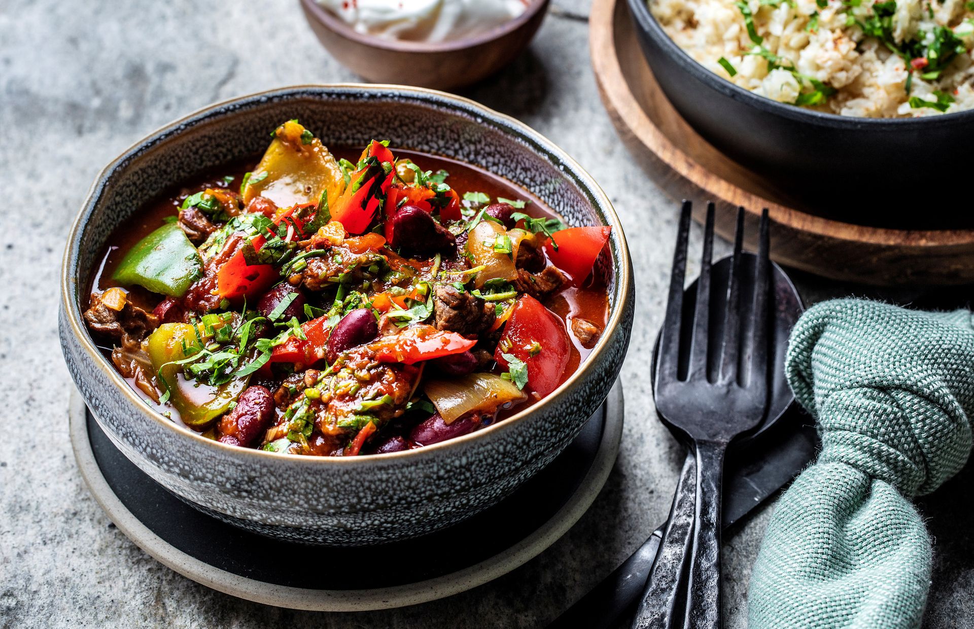 Mørk keramikkbolle fylt med en fargerik grønnsaksgryte med røde bønner, paprika, squash og urter i kraftig saus, fotografert ovenfra. Ved siden av står en svart gaffel og et grønt tekstil på grå steinaktig underlag.