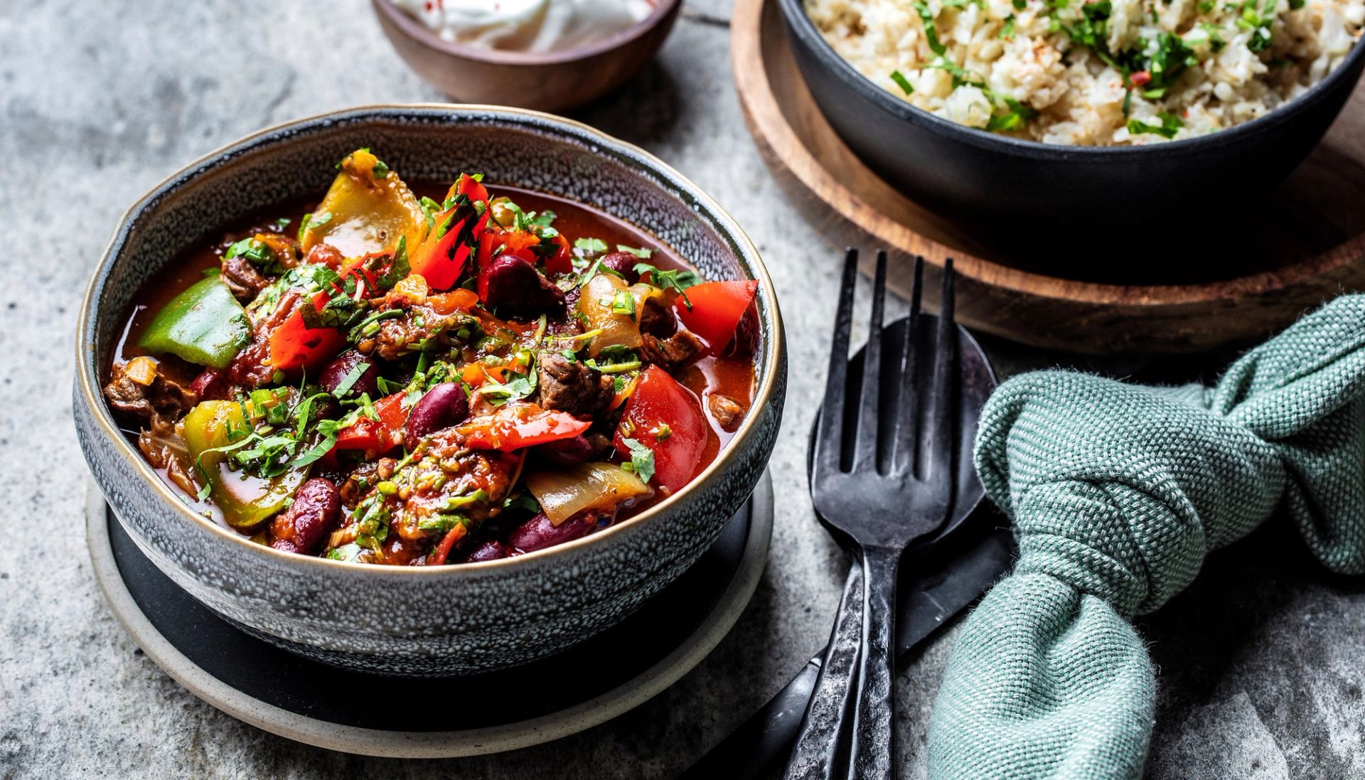 Mørk keramikkbolle fylt med en fargerik grønnsaksgryte med røde bønner, paprika, squash og urter i kraftig saus, fotografert ovenfra. Ved siden av står en svart gaffel og et grønt tekstil på grå steinaktig underlag.