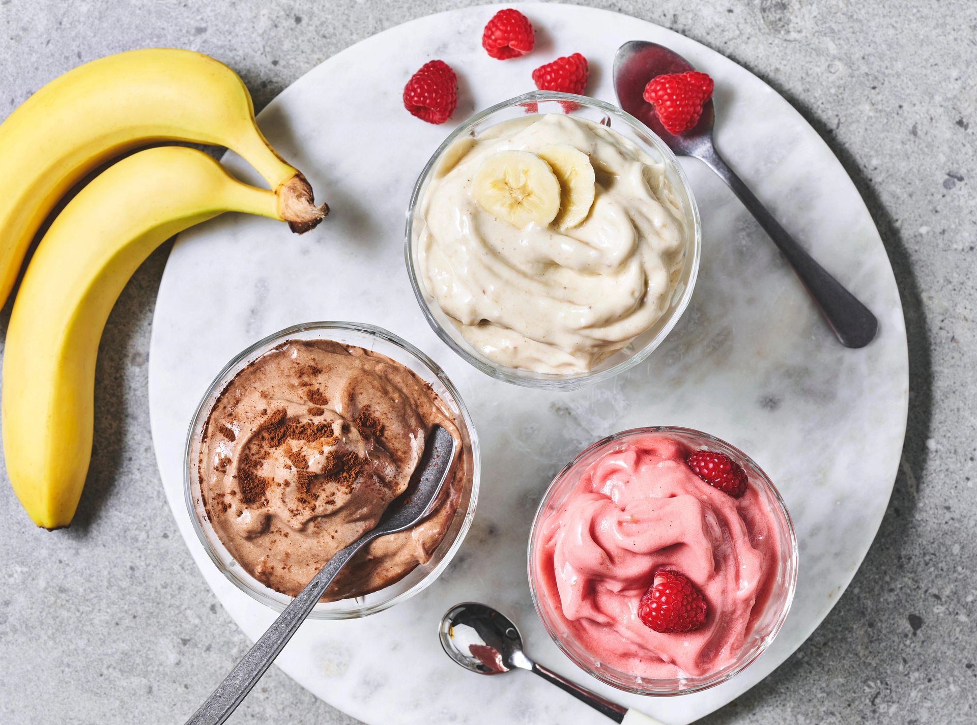 Kremet iskrem av banan i tre skåler, en rosa, en hvit og en brun. Skålene er plassert på et marmorbrett og det ligger bringebær strødd. Ved siden av ligger to bananer.