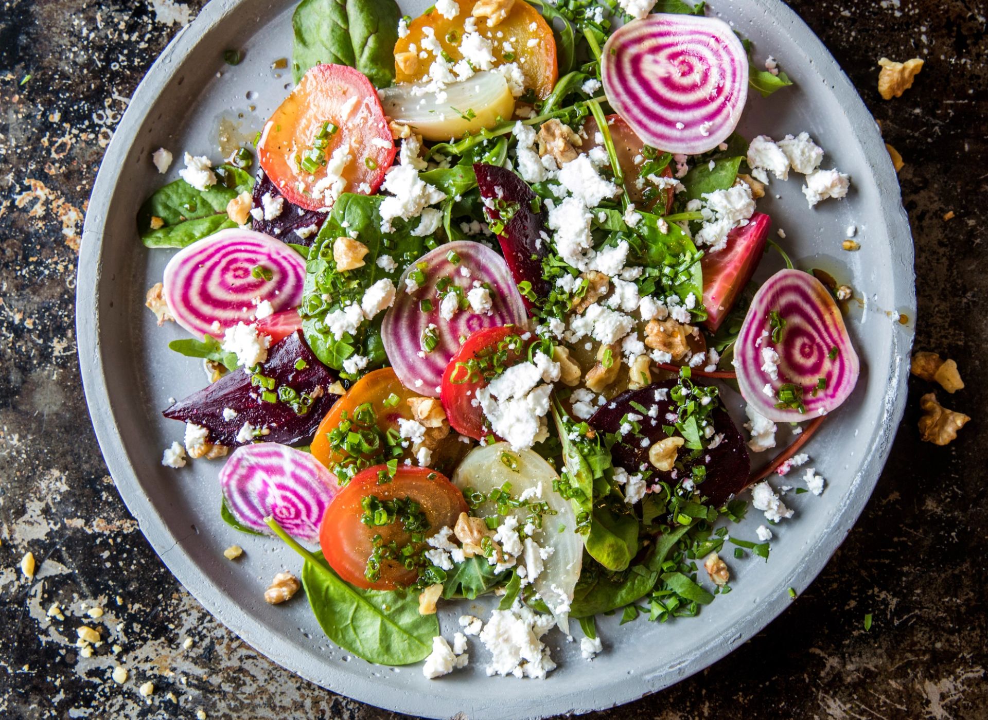 Fargerik salat servert på en grå tallerken med skiver av rødbeter i gule og lilla nyanser, spinatblader og ruccula, toppet med smuldret hvit ost, hakkede urter og biter av valnøtter.