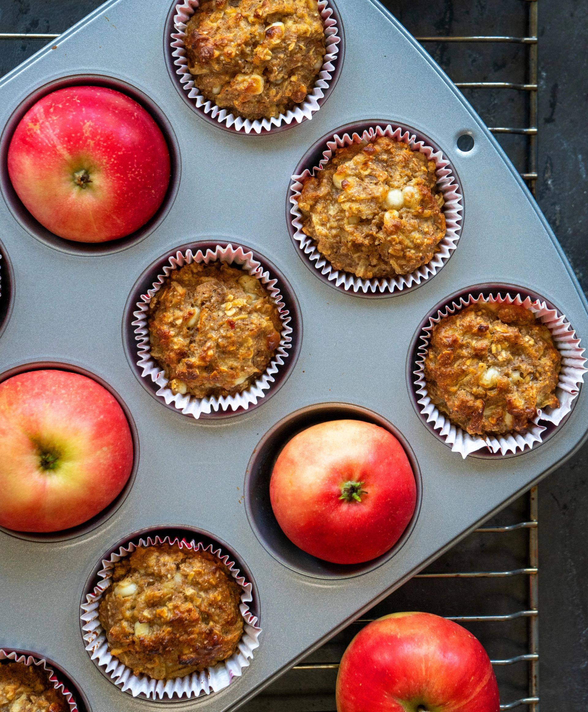 Et muffinsbrett med nybakte eplemuffins i papirformer og hele røde epler plassert i noen av rommene, sett ovenfra på en rist
