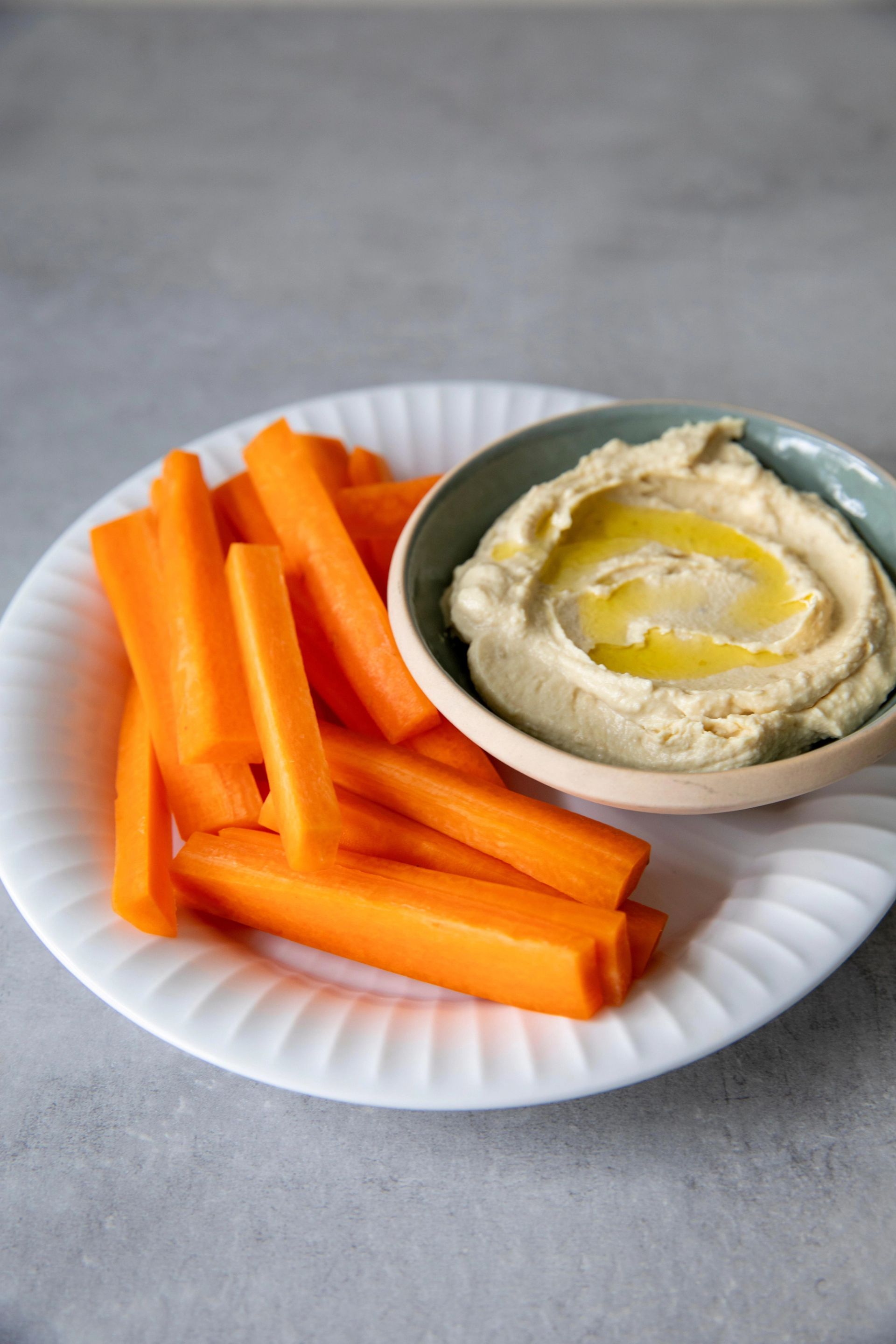 Gulrotstaver servert på en hvit tallerken ved siden av en liten skål med kremet hummus med olivenolje på toppen.