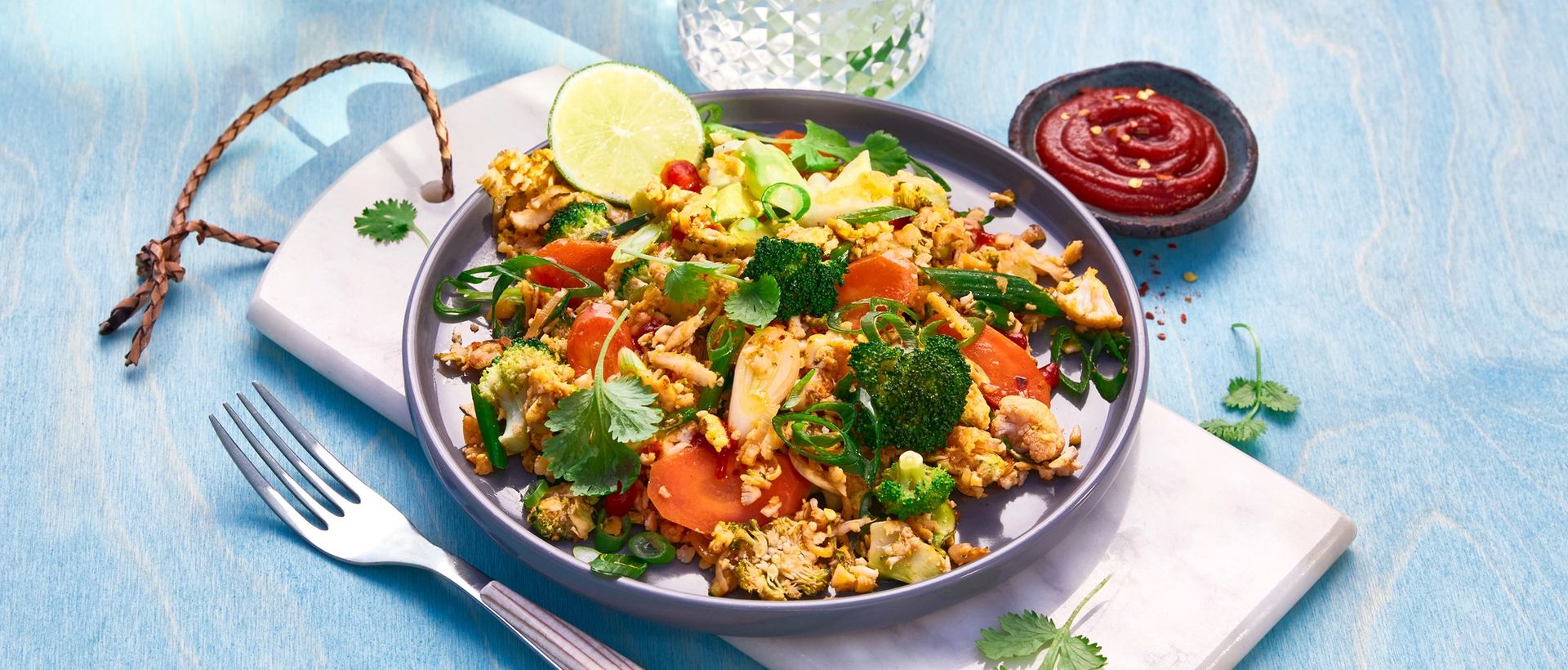 Blomkålris med stekt egg, grønnsaker og frisk koriander servert på tallerken med lime og chilisaus