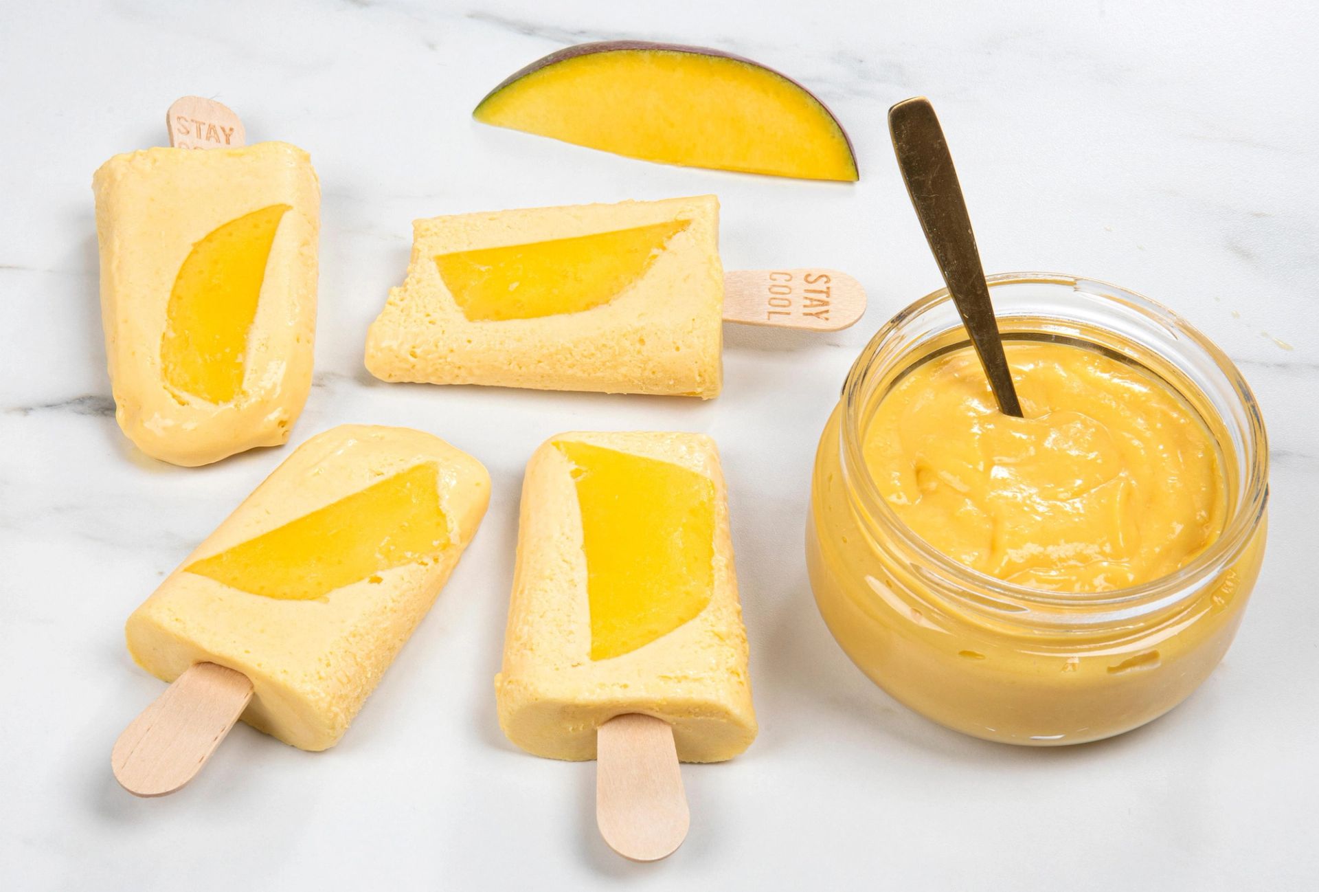 Fire ispinner og en glasskrukke med kremet mangoblanding står på et lyst marmorbord, alle med synlige biter av gul mango.