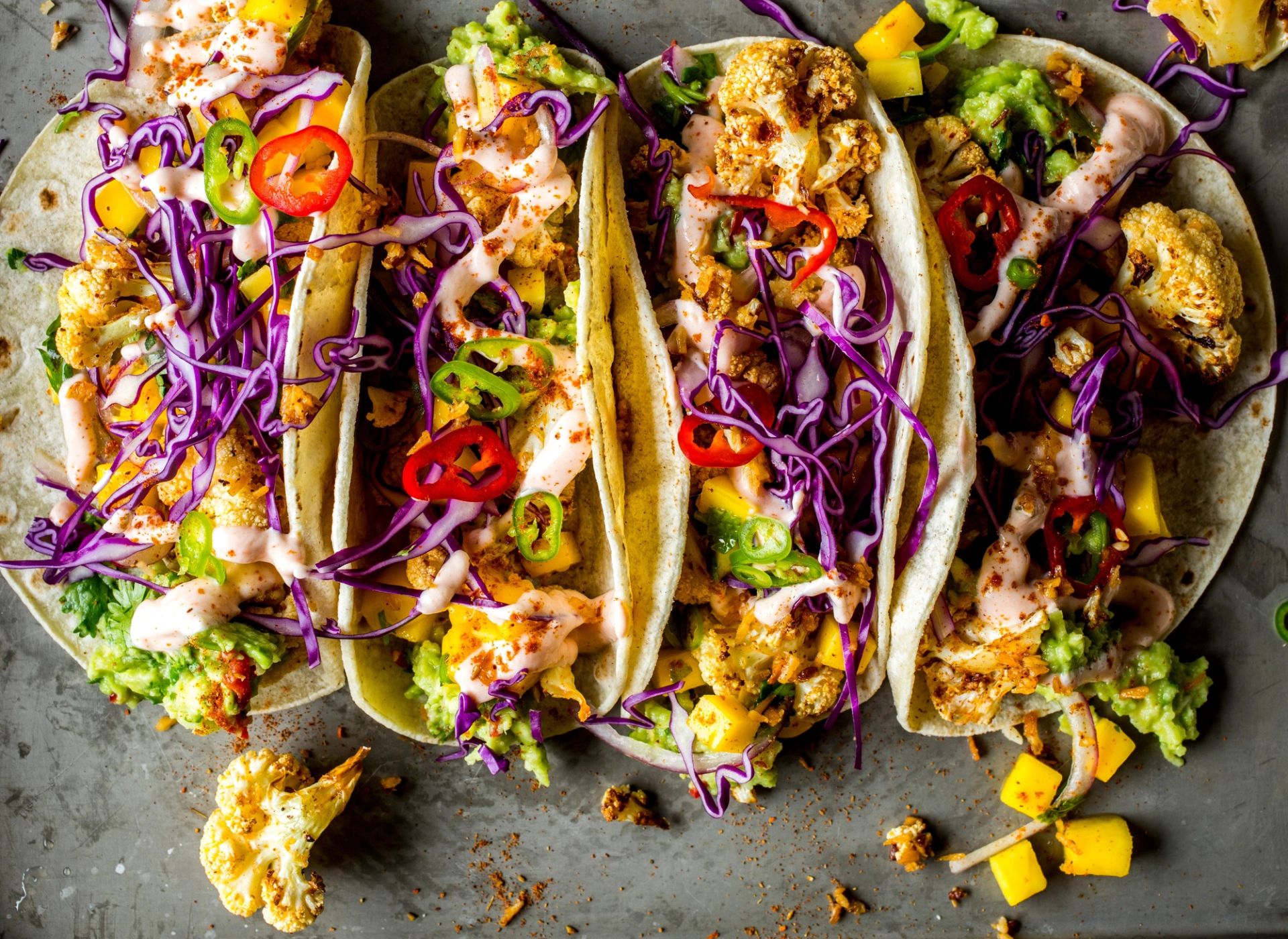 Oversiktsbilde av fire myke tacos fylt med ovnsbakt blomkål, lilla rødkål, quacamole, mangobiter og skiver av rød og grønn chili, toppet med en lys saus.