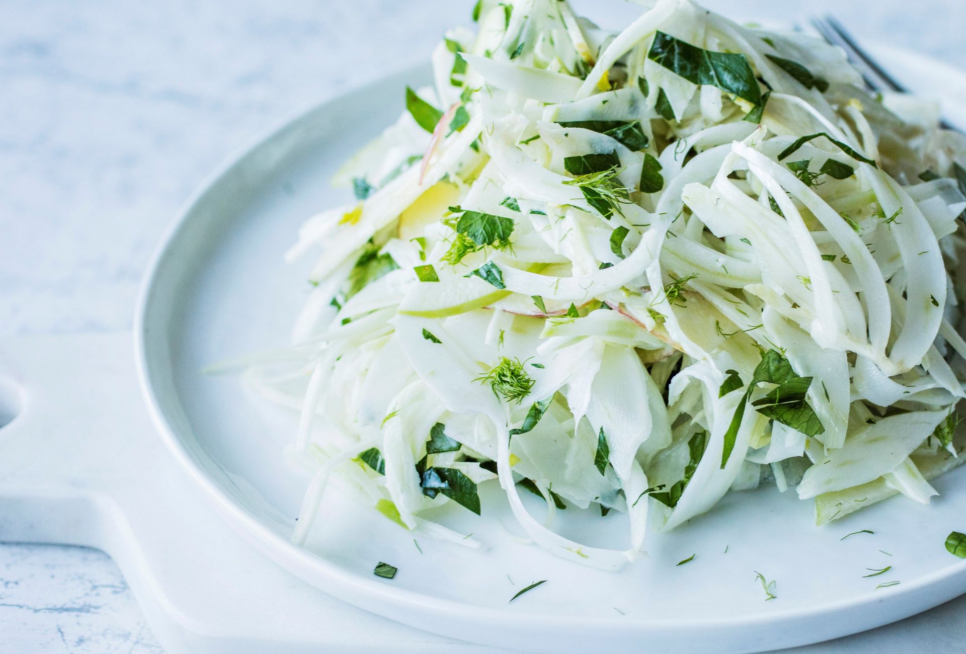 Tynt strimlet coleslaw med brokkoli, fennikel og eple servert på en lys tallerken, med friske urter fordelt over.