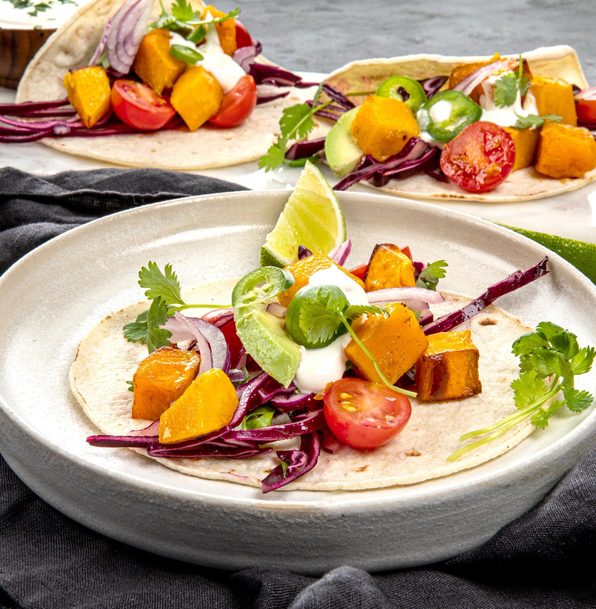 Nært bilde av en myk tortillalefse fylt med ovnsbakte gresskarbiter, rødkålstrimler, cherrytomater, avokado, jalapénosskiver, rød løk, frisk koriander og en klatt rømme. En limebåt ligger på toppen. I bakgrunnen står to lignende tacos på et grått underlag.