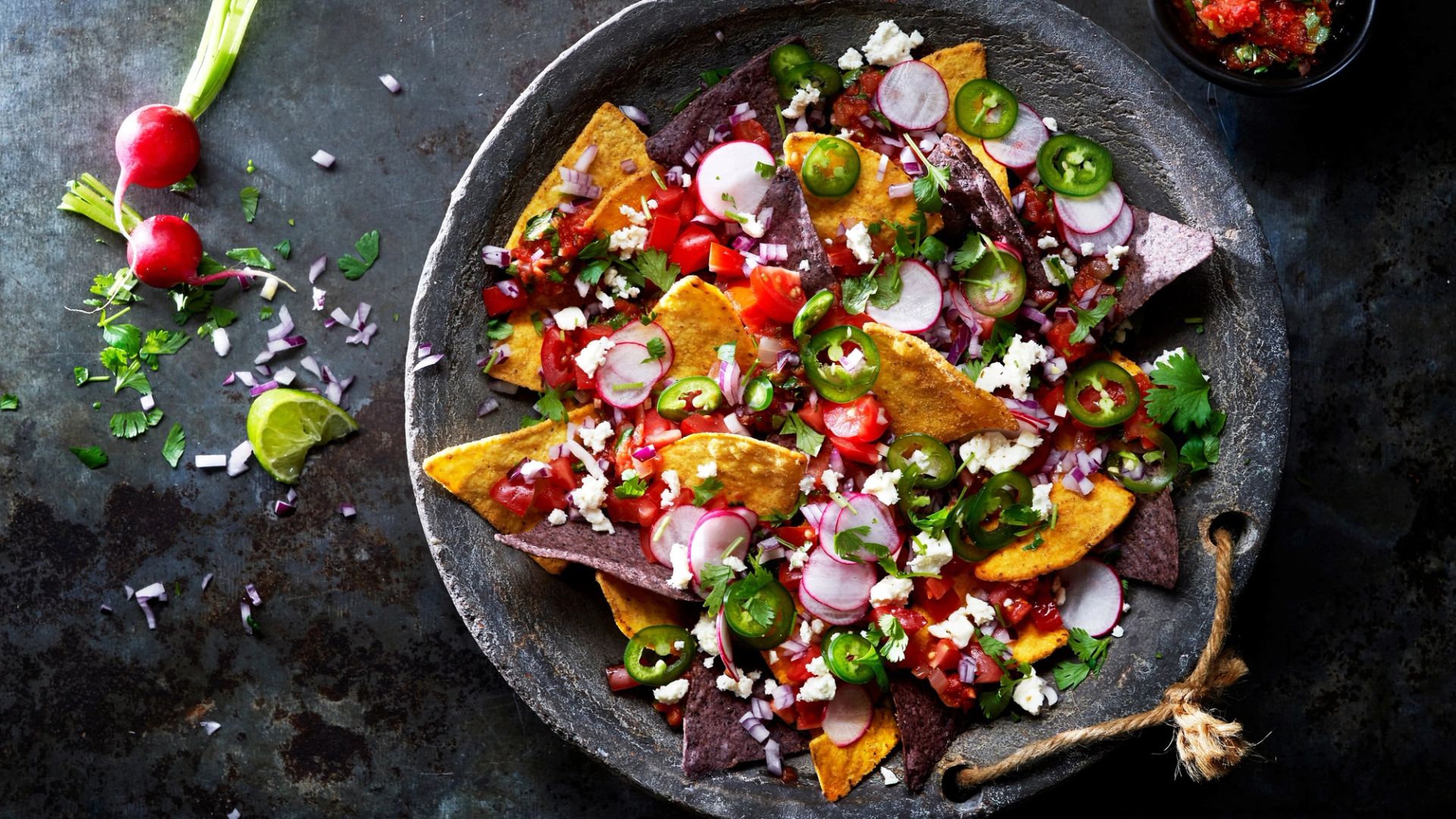 En steintallerken med nachos av blå og gule maischips, toppet med skivede reddiker, jalapeño, tomater, smuldret ost og frisk koriander. Tallerkenen står på et mørkt bord med lime og reddiker ved siden av.