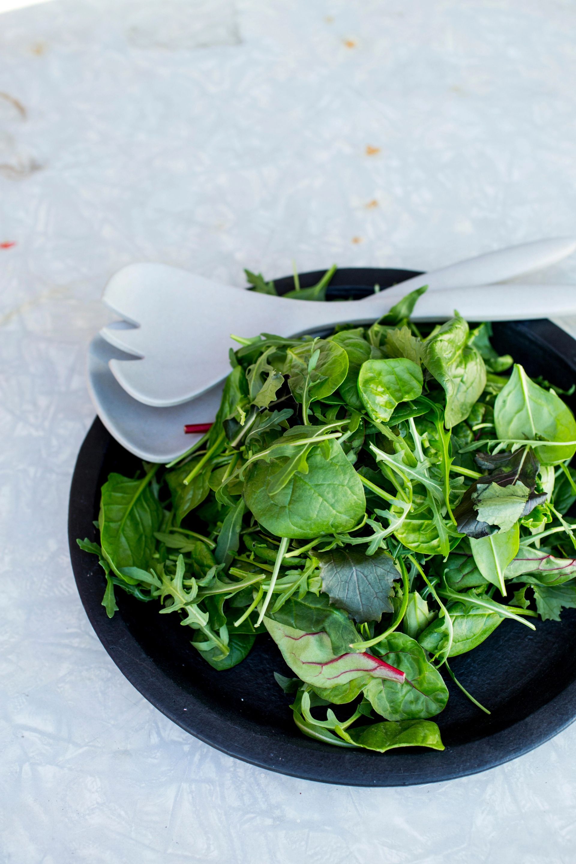 Skål med blandet grønn salat servert i mørk bolle, med lyse og mørke blader, sett ovenfra, med hvitt salatbestikk ved siden av.