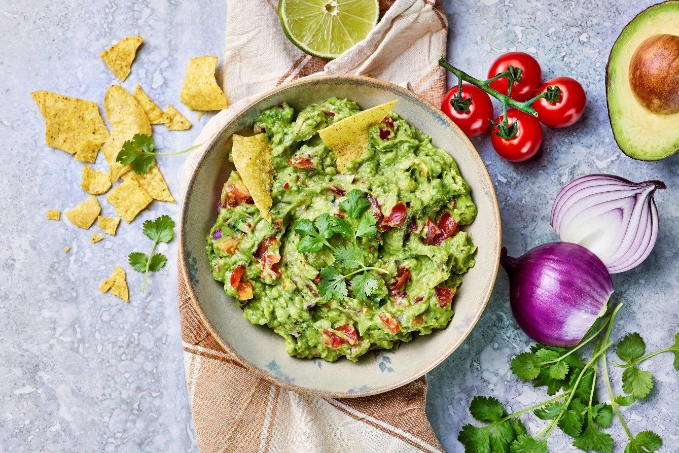 Bolle med hjemmelaget guacamole laget av avokado, chili og koriander, servert med tortillachips, lime, cherrytomater og frisk avokado rundt på bordet.