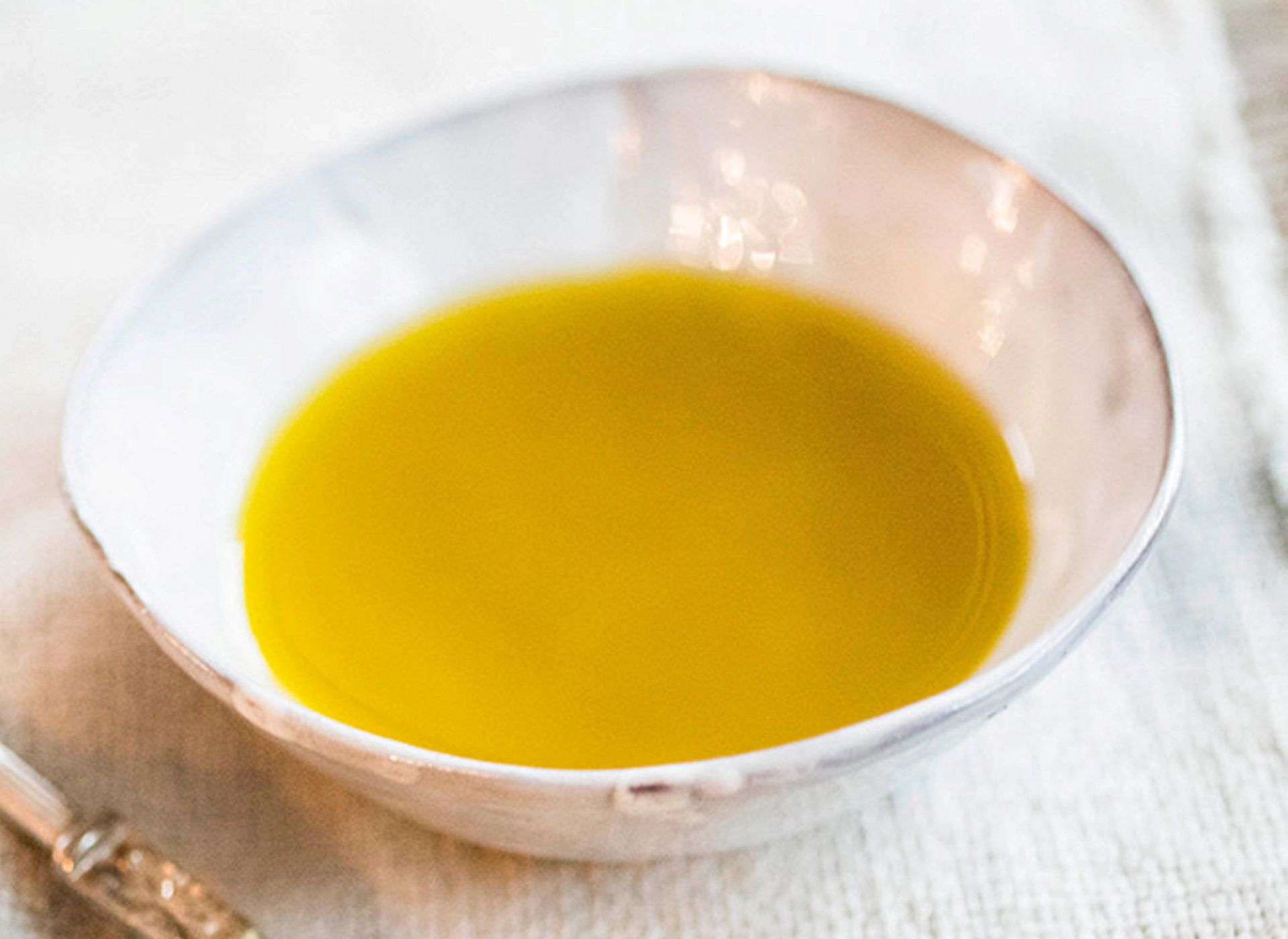 En skål med gyllengul dressing servert på et lyst tekstilunderlag. Den glatte overflaten og den klare fargen fremheves av mykt naturlig lys, mens en liten skje ligger delvis synlig ved siden av skålen.