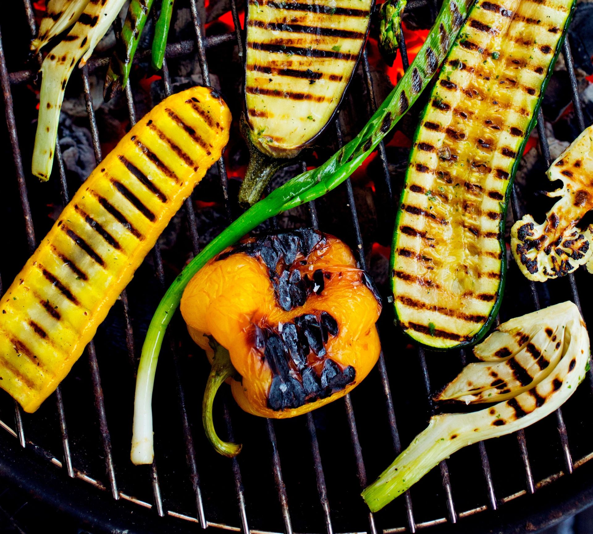 Grillede grønnsaker ligger på en grillrist over glødende kull. På risten ligger blant annet skiver av squash med grillstriper, asparges, en halv rød paprika med svidde kanter, og biter av rødløk og hvitløk.