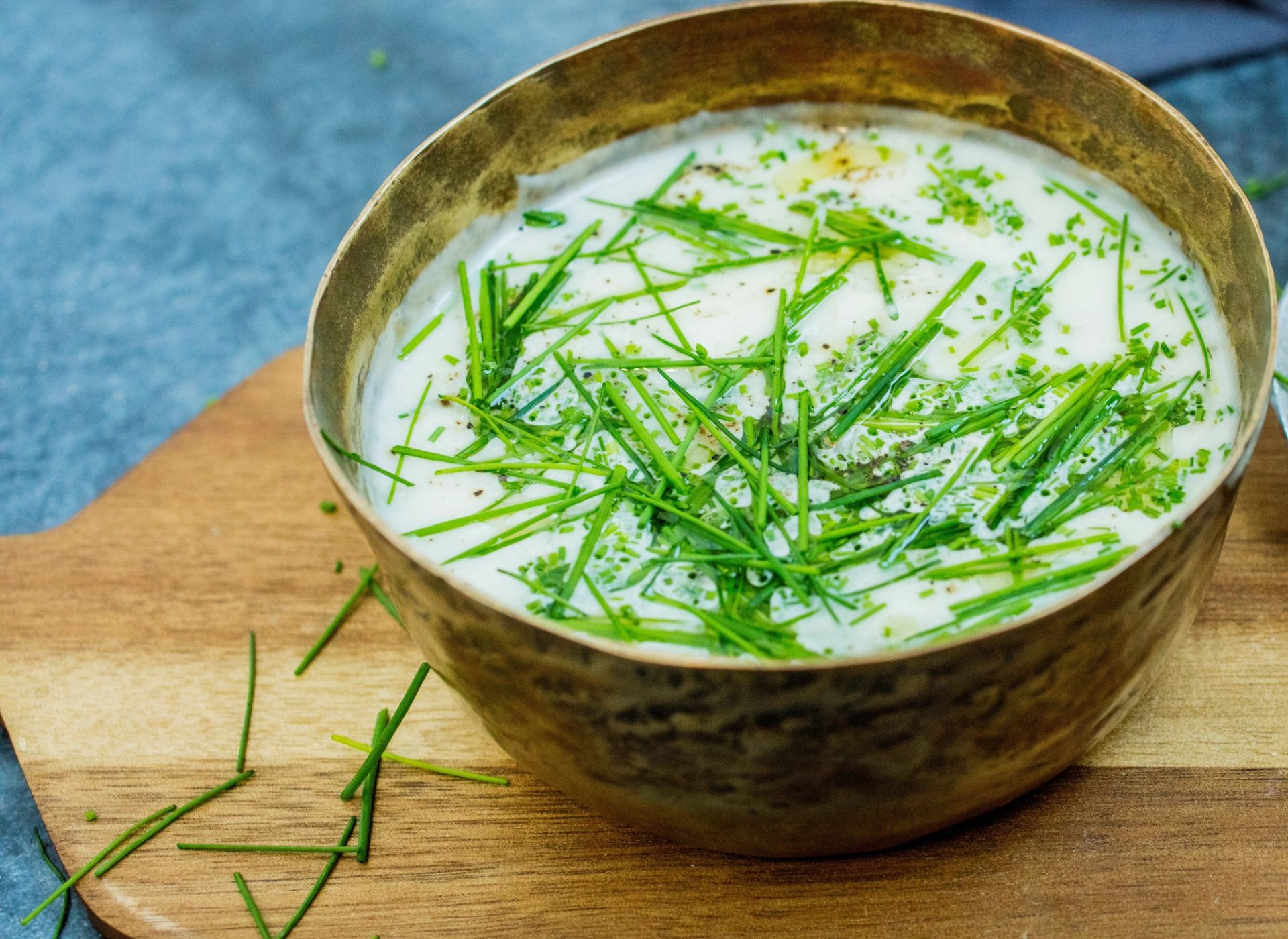 Hvit kremet suppe i en kobberfarget skål, toppet med rikelig med finhakket gressløk, servert på en trefjøl på blå-grått underlag.