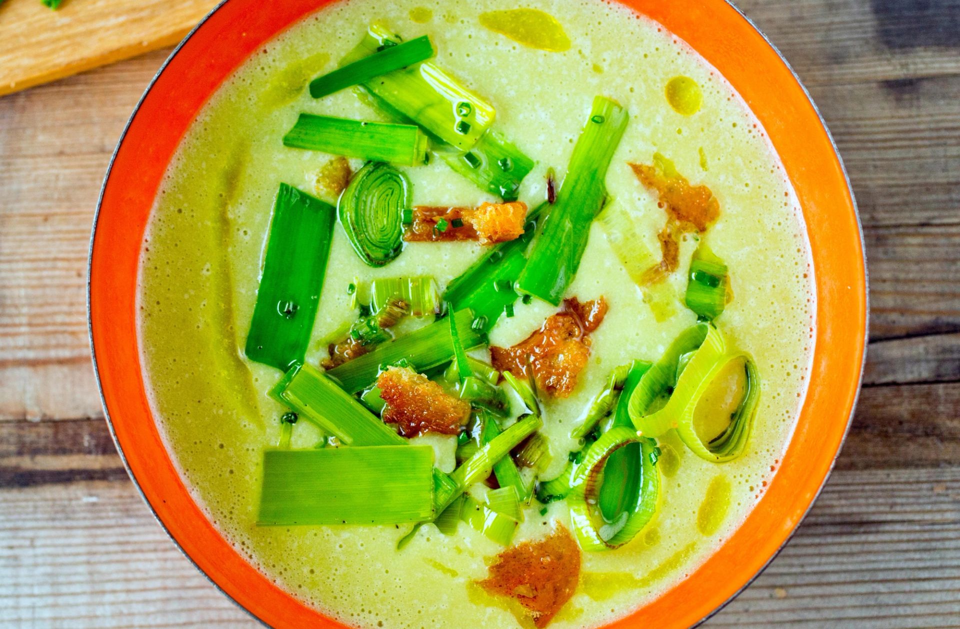En skål med kremet purre- og potetsuppe, toppet med biter av sprøstekt purre og små gyllenbrune brødkrutonger. Suppen er lysegrønn og står på et rustikt trebord. I bakgrunnen ligger en fjøl med hakket purre.
