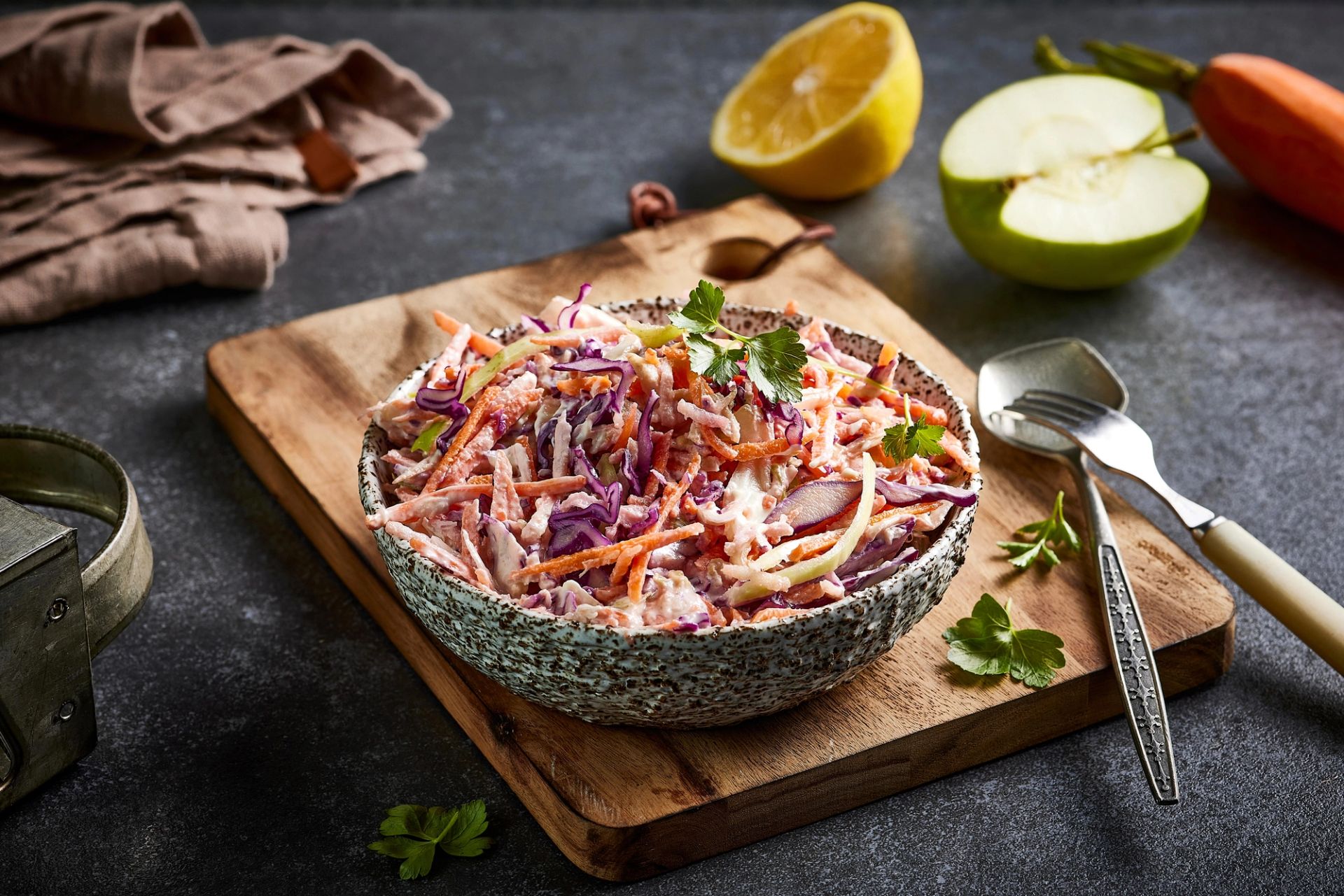 Skål med kremet råkostsalat av strimlet kål, gulrot og eple, toppet med friske urter, plassert på et trebord.