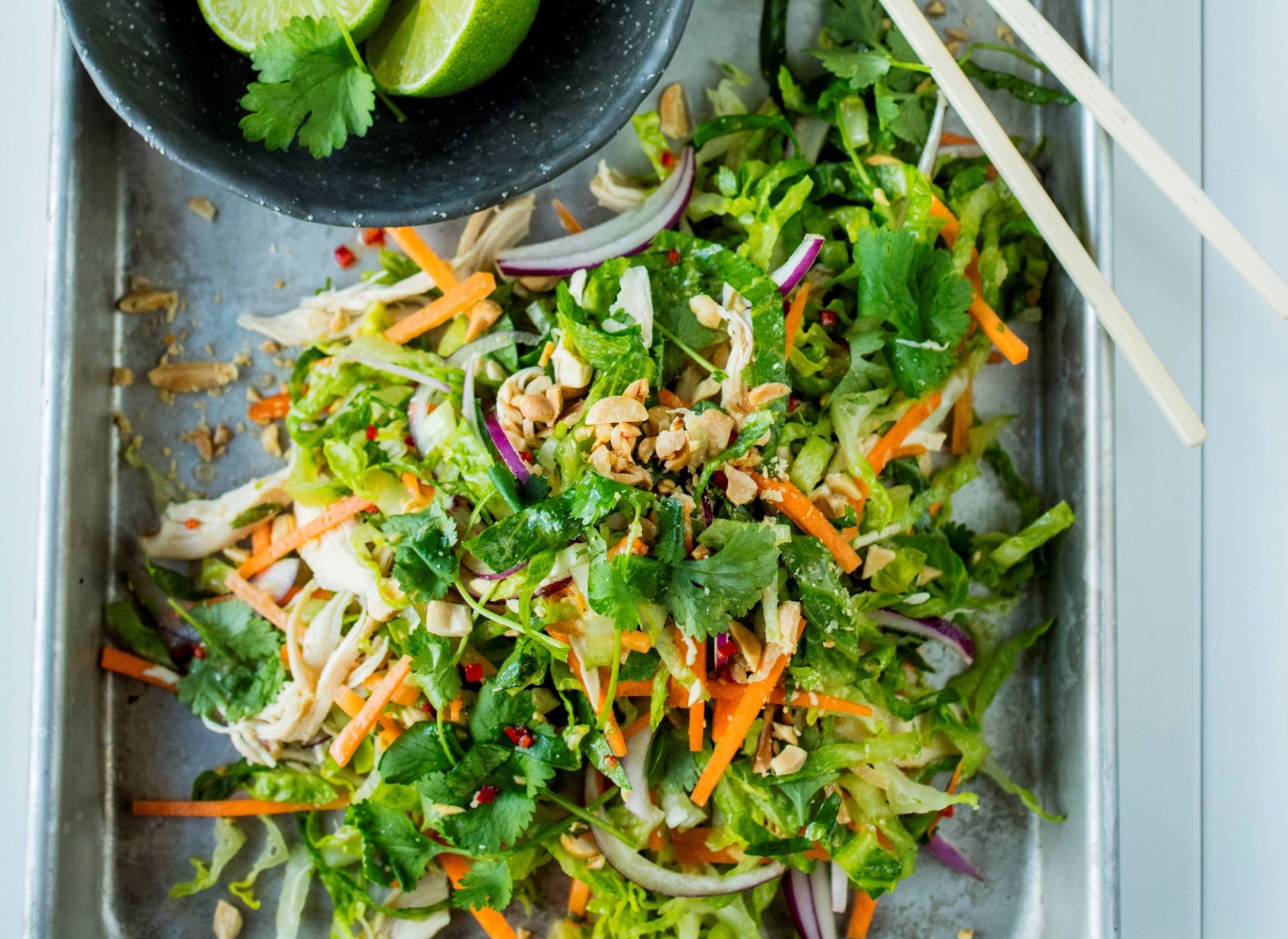 Frisk salat med hjertesalat, strimlet kylling, gulrot, rødløk, koriander og hakkede peanøtter, servert på et bakebrett. Toppet med lime og asiatisk‑inspirert dressing.