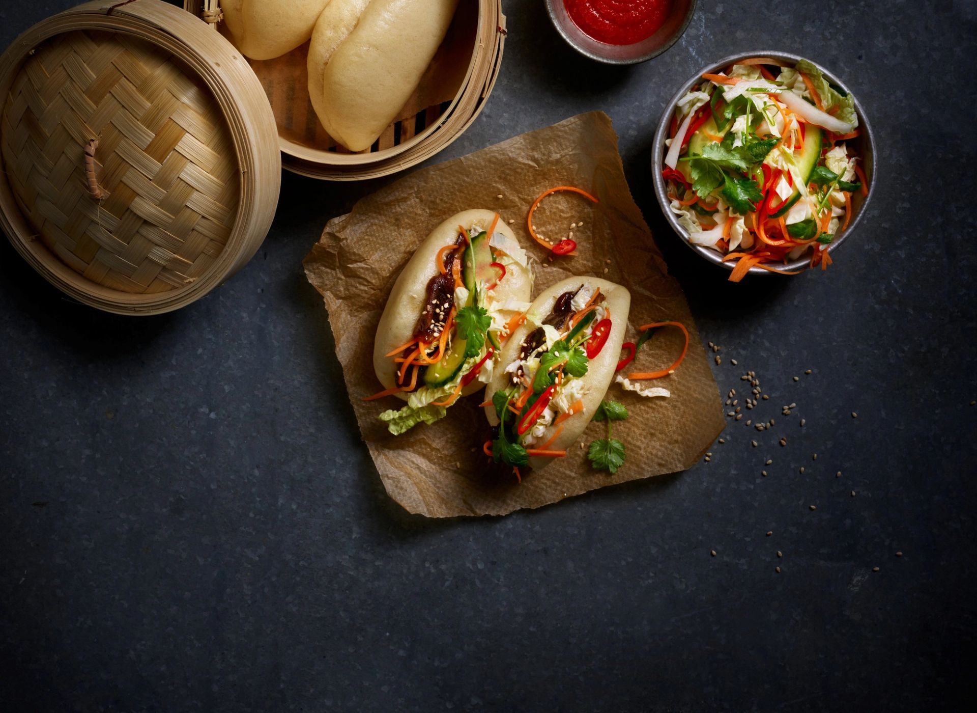 Steamed buns (bao) med sopp, sprø grønnsaker og sursøt salat servert på bakepapir med bambusdamper og chilisaus.