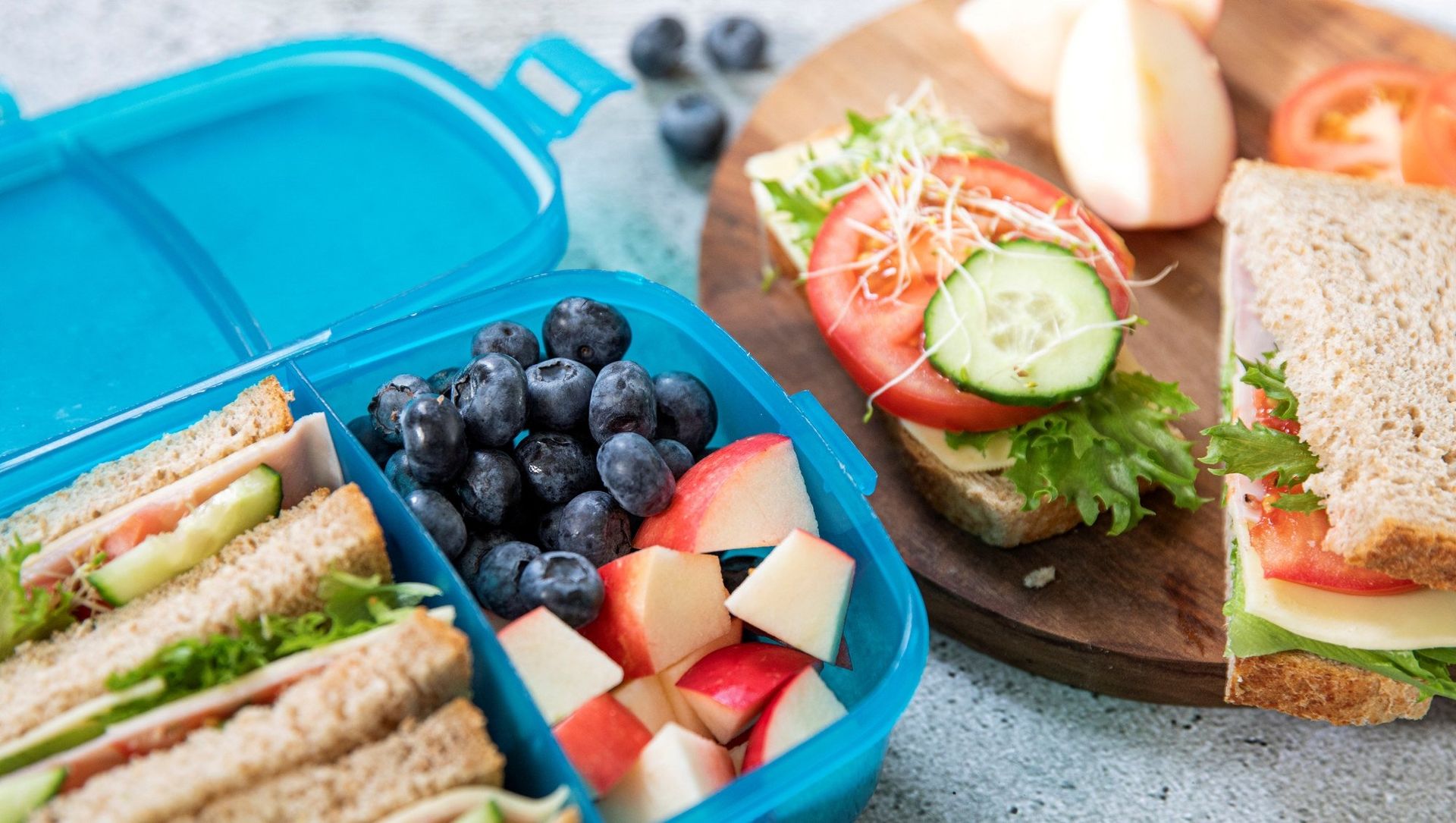 Matboks med grove sandwiches, blåbær og oppskårne eplebiter ligger åpen på et bord. Ved siden av matboksen står åpne brødskiver toppet med salat, agurk, tomat og spirer, samt et halvt eple og noen blåbær.