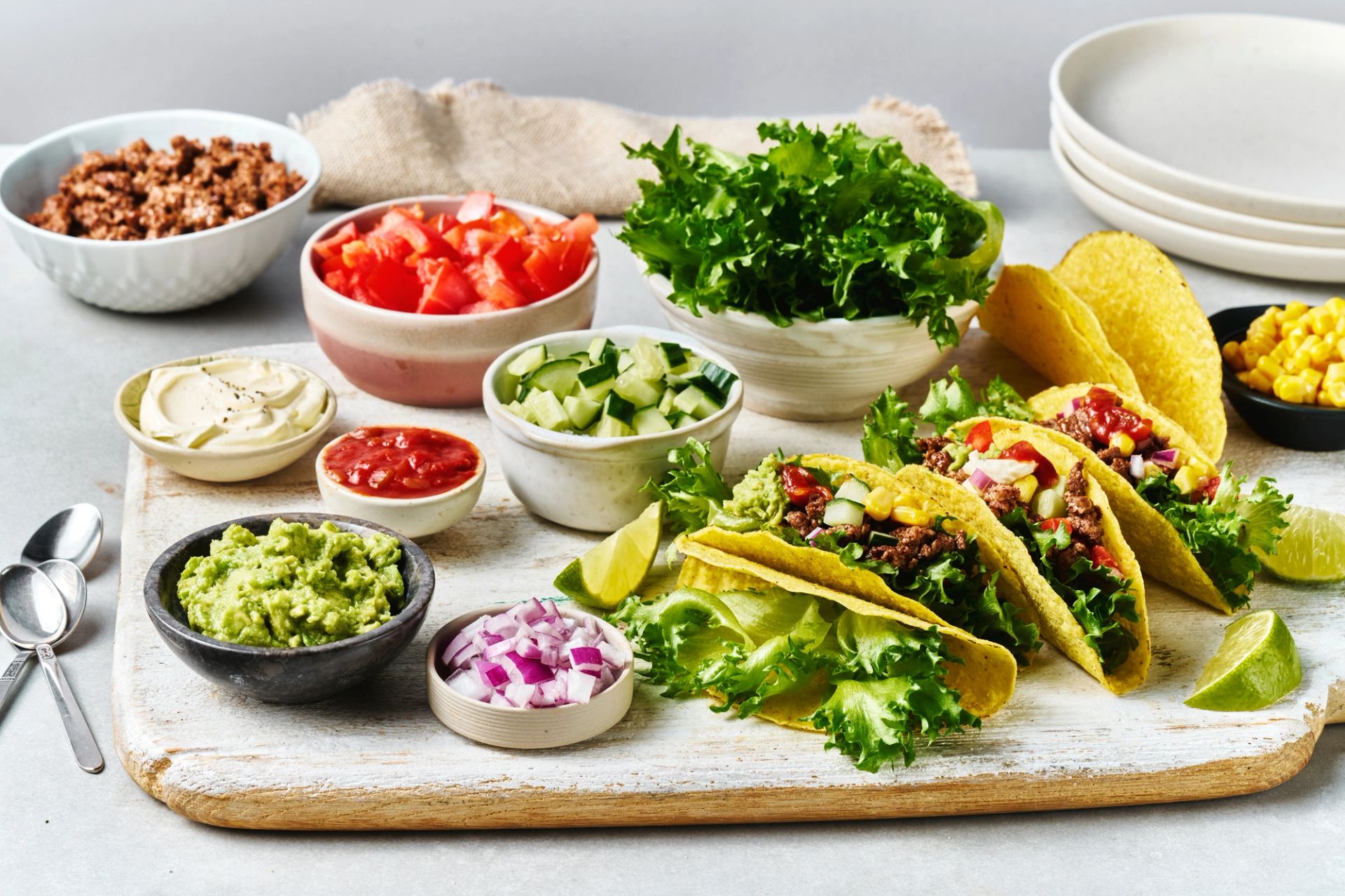 En stor, hvitmalt trefjøl fylt med alt som trengs til et tacomåltid. Til høyre står fire sprø tacoskjell fylt med kjøttdeig, salat, mais og salsa. Rundt på fjøla er det plassert små skåler med tilbehør: kremet guacamole, finhakket rødløk, terninger av agurk og tomat, revet salat, tacokjøttdeig, rømme, salsa og mais. Noen båter med frisk lime ligger spredt mellom skålene.