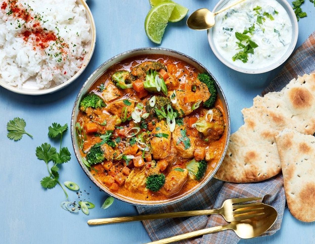 Et fargerikt matbilde sett ovenfra viser en skål med kremet, oransje curry fylt med biter av grønnsaker som brokkoli og paprika, toppet med frisk koriander og finhakket vårløk. Rundt skålen ligger tilbehør: en bolle med hvit ris drysset med krydder, et lite fat med yoghurtdressing med urter, limebåter, samt naanbrød. Bestikk i gullfarge og noen løse urter er plassert dekorativt på en lys blå bakgrunn.
