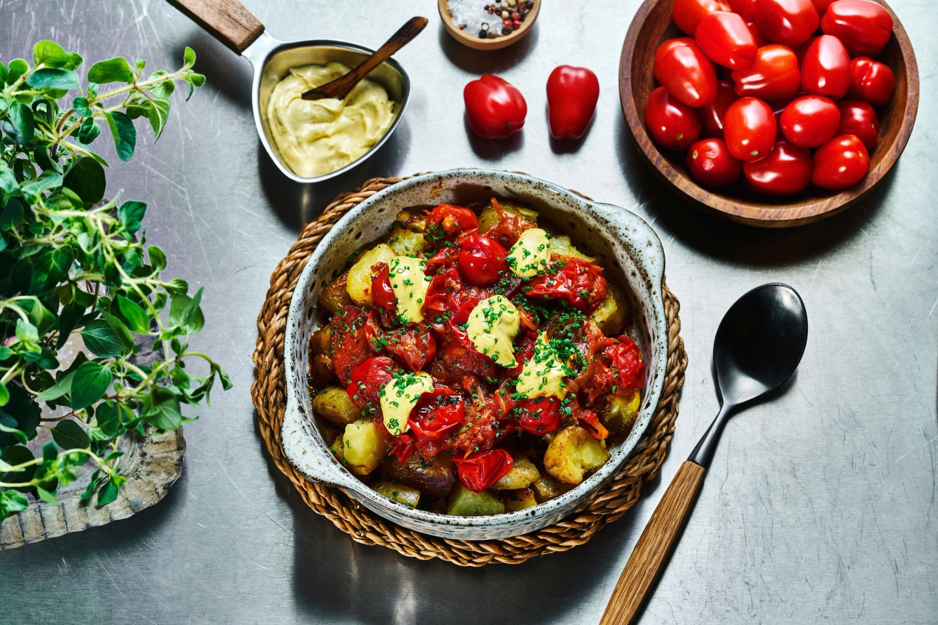 En ildfast form sett ovenfra med ovnsbakte poteter toppet med tomater, chorizopølse og friske urter, omgitt av en skål med cherrytomater, en skje og aioli.