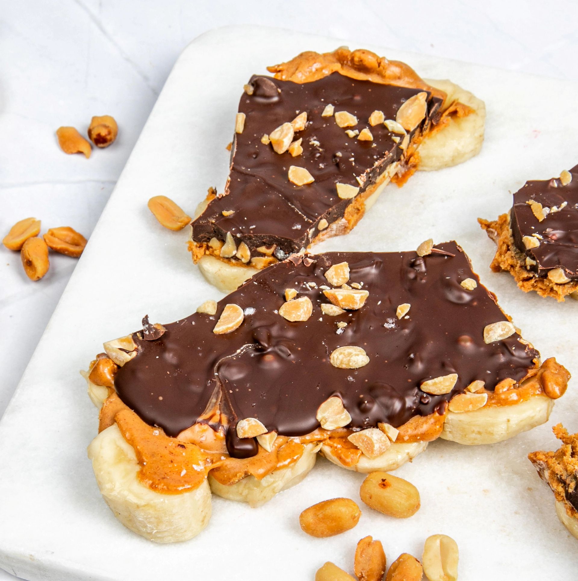En snackbar laget av skiver med banan toppet med peanøttsmør, et lag mørk sjokolade og hakkede peanøtter. Barene ligger på en lys marmorlignende overflate med noen løse peanøtter rundt.