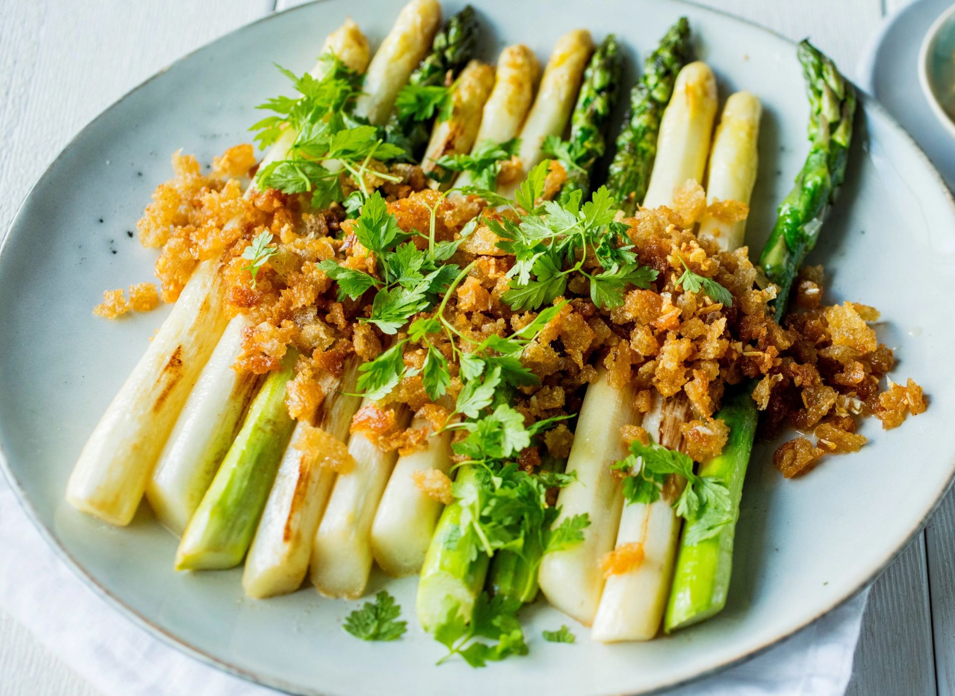Stekte grønne og hvite asparges med sprøtt dryss og friske urter, servert på lys tallerken med gyllen og crispy topping
